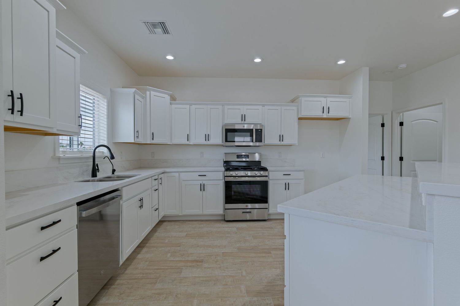 Modern white kitchen with stainless steel appliances, quartz island, recessed lighting, and shaker cabinets on light tile floors.