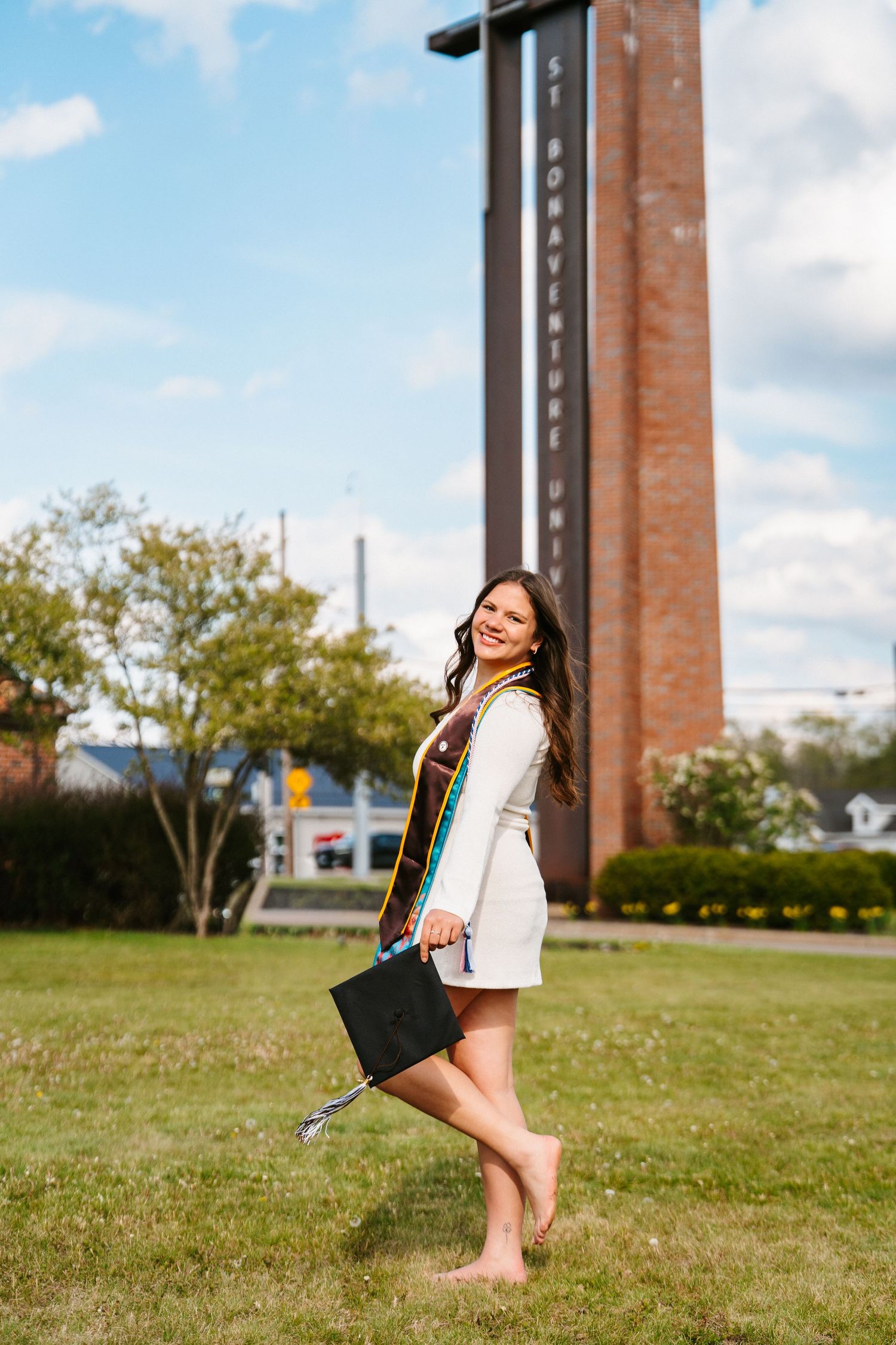 Barefoot graduate in a white jacket and honor stole holds her cap while posing on grass near a tall brick tower sign.