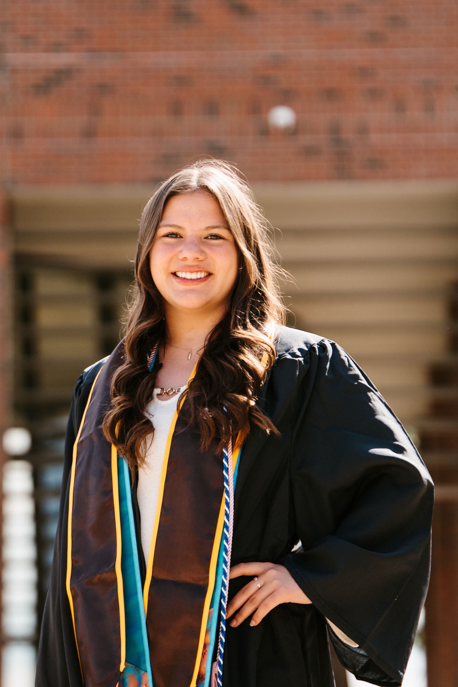 Smiling graduate with long wavy hair wears a black gown and teal and gold stoles while posing outside a red brick building.