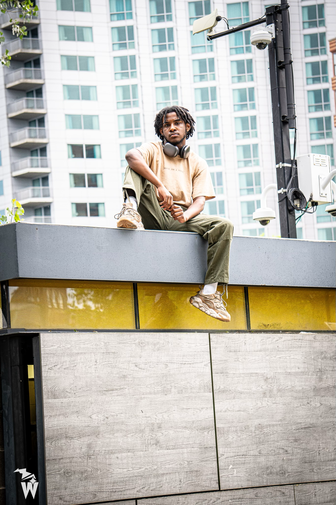 Young man wearing headphones sits on a gray and yellow ledge, looking at the camera, with a high-rise building in the background.