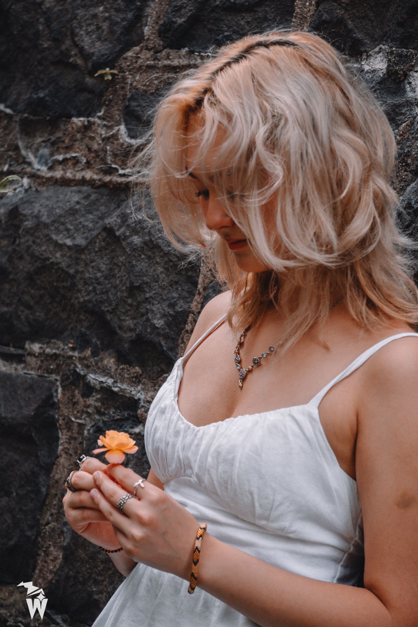 A woman with wavy blonde hair in a white sundress looks down at a small orange flower while standing before a dark stone wall.