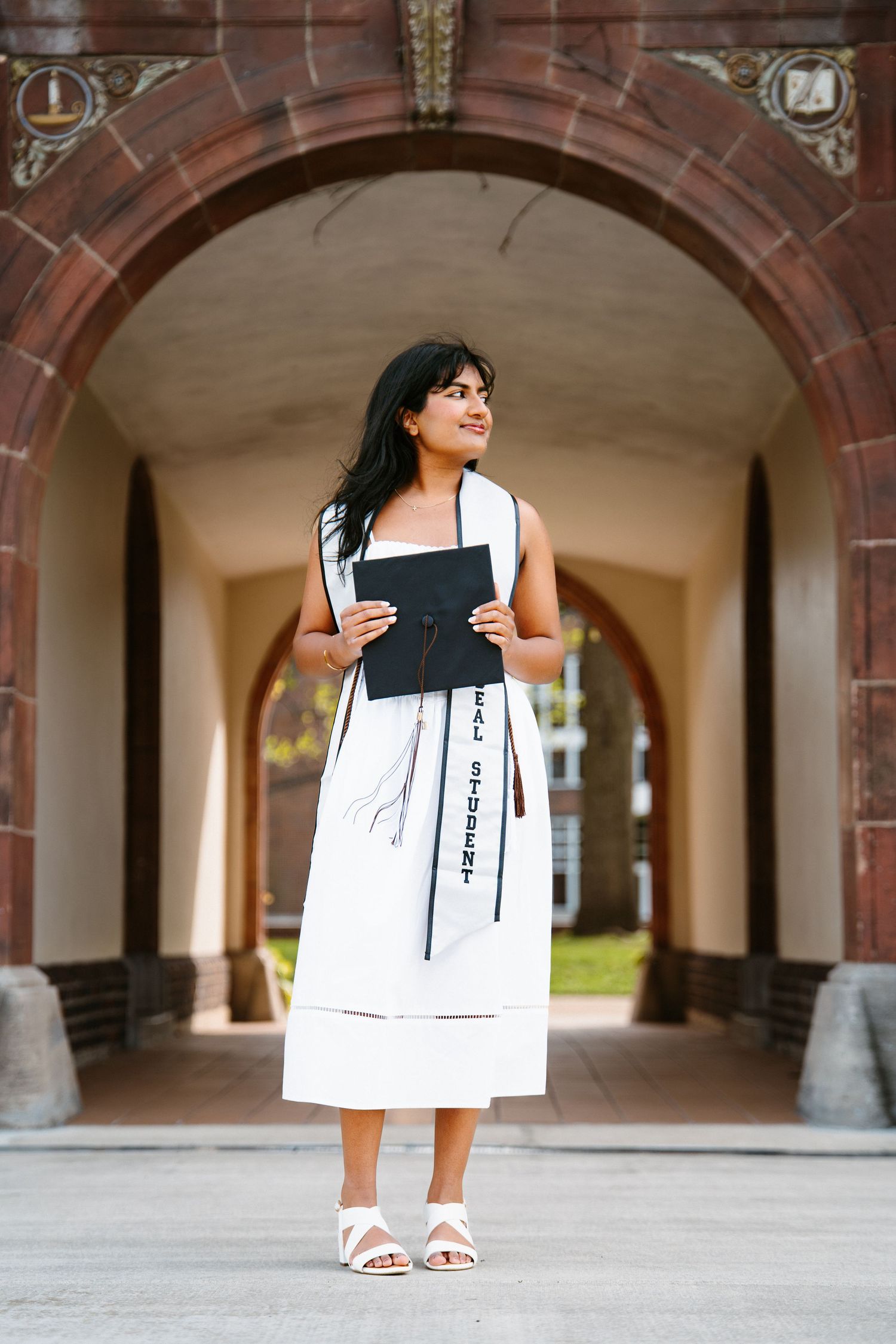 A graduate in a white dress and Student Athlete stole holds her black cap and looks aside while standing in a brick archway.