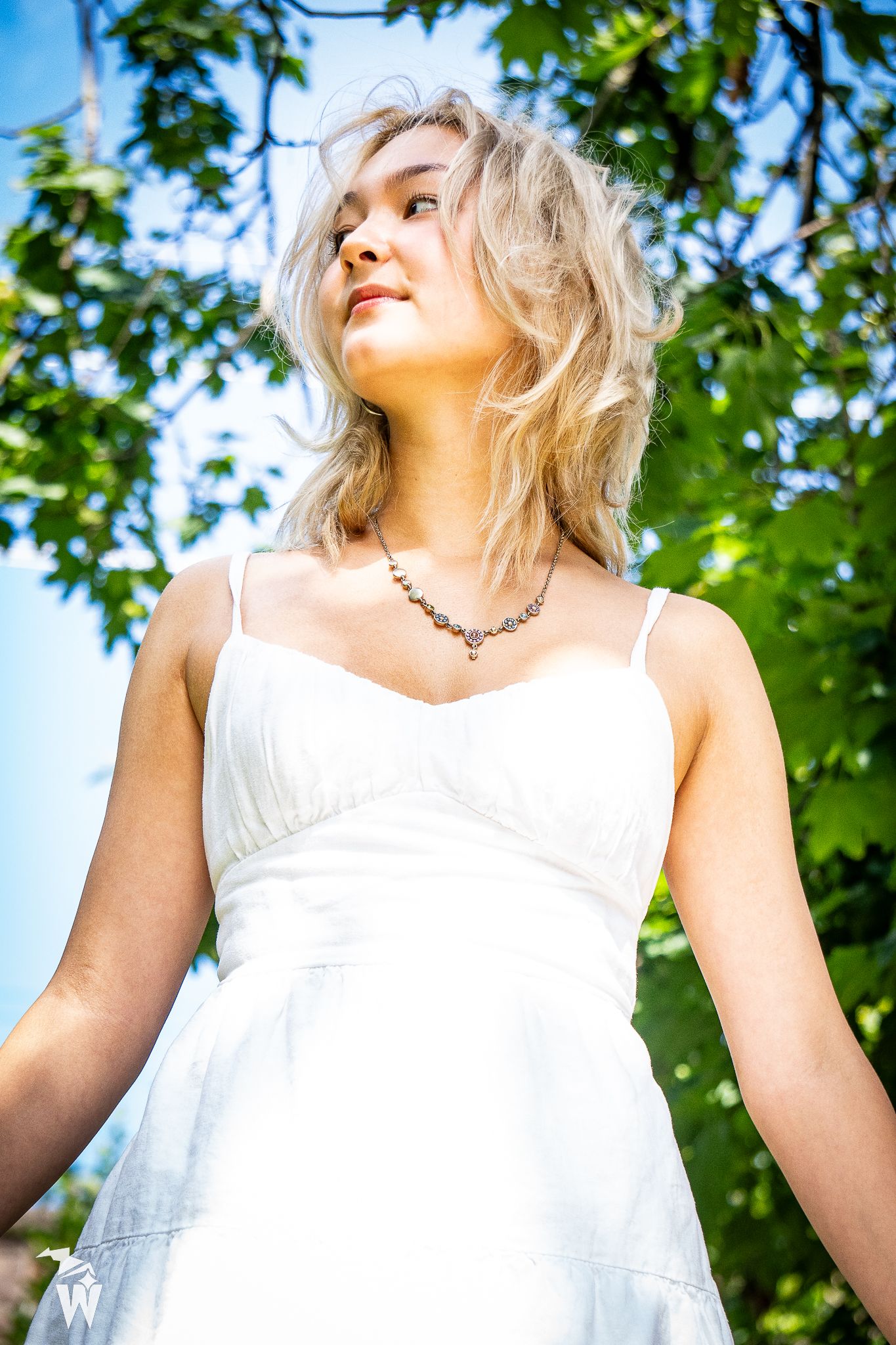 Low-angle portrait of a blonde woman in a white dress and beaded necklace gazing upward beneath green trees and blue sky.