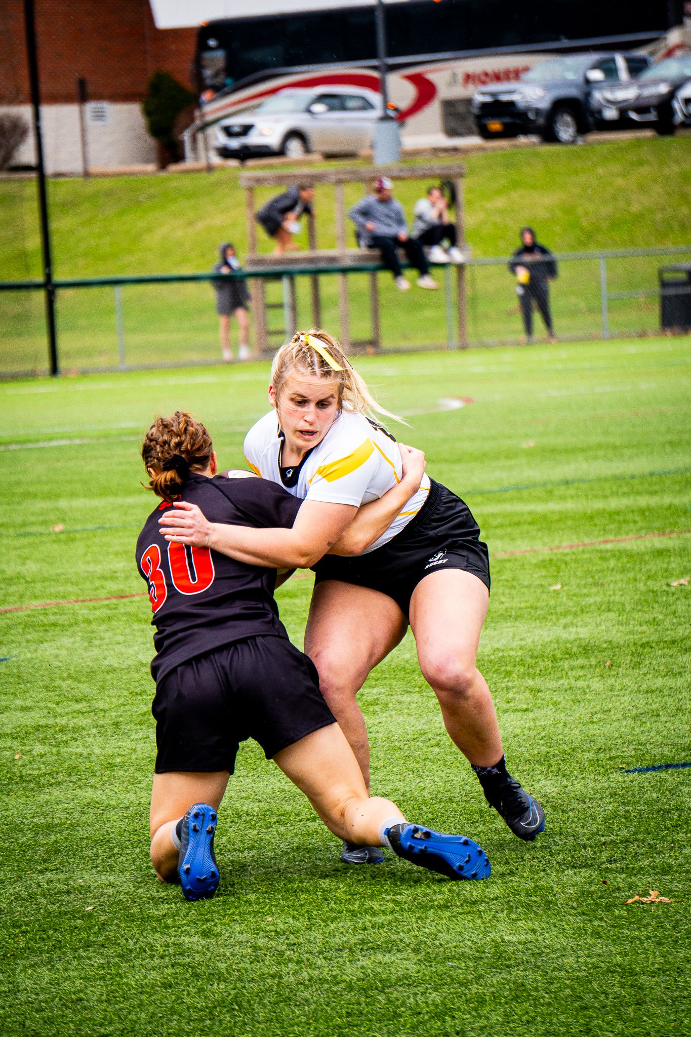A women's rugby player in a white and gold jersey wraps up a black-jerseyed number 30 in a tackle while fans watch from the sidelines.