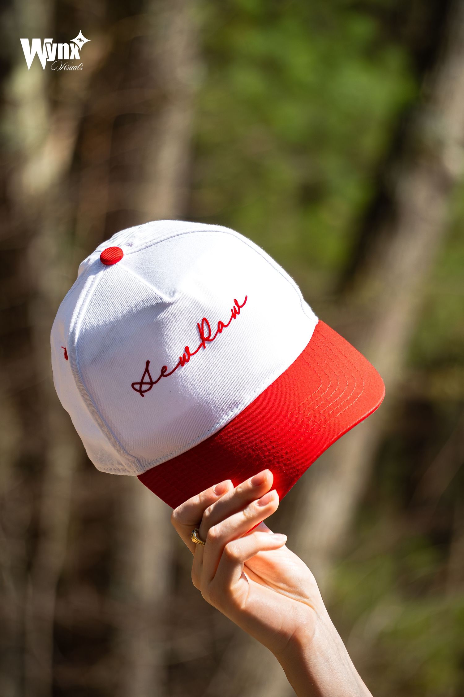 Hand holds up a white and red Sewlaw snapback cap with red script embroidery against a blurred sunlit forest background.
