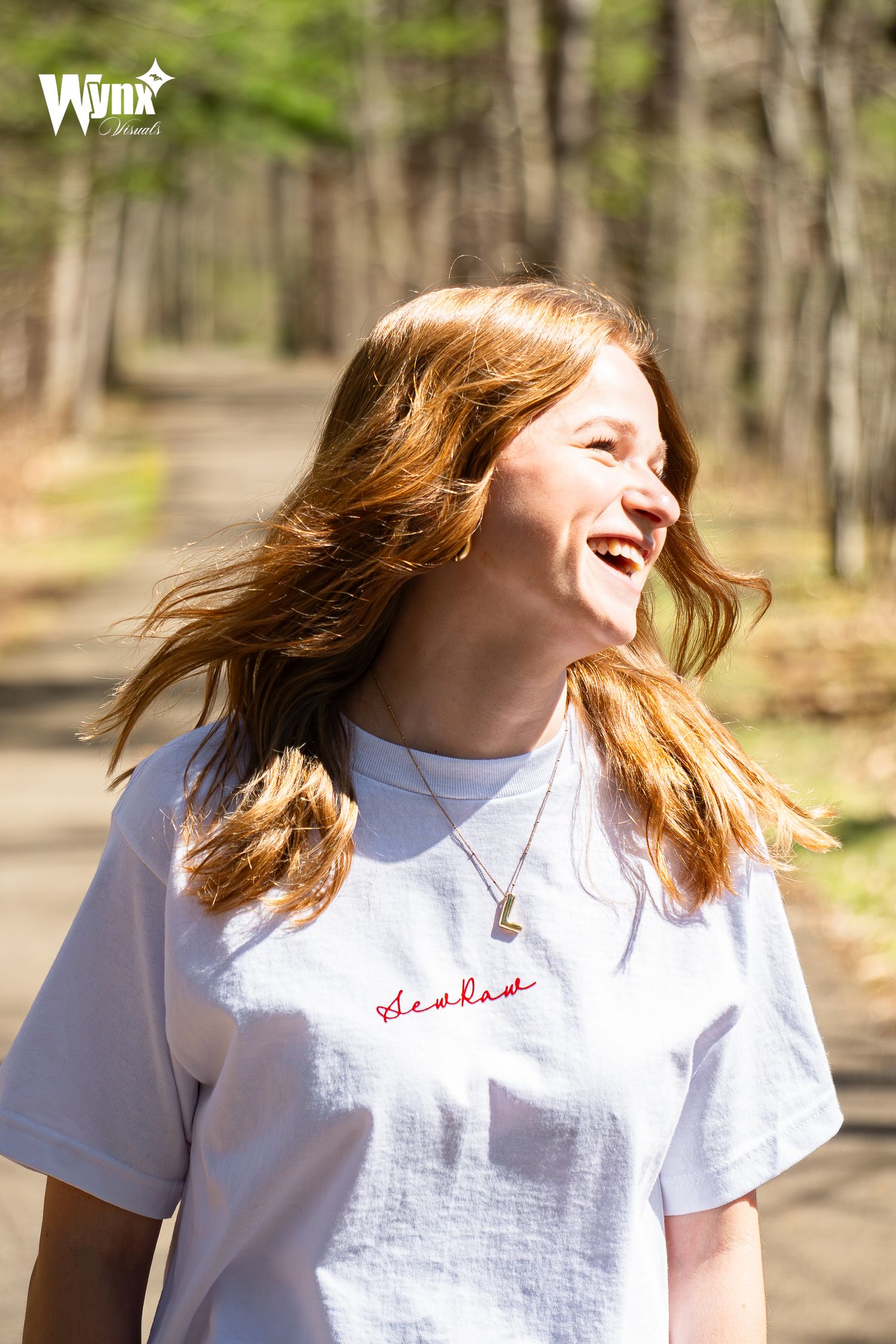 Hand holding up a white and red embroidered baseball cap with a red button against a blurred sunlit forest background.
