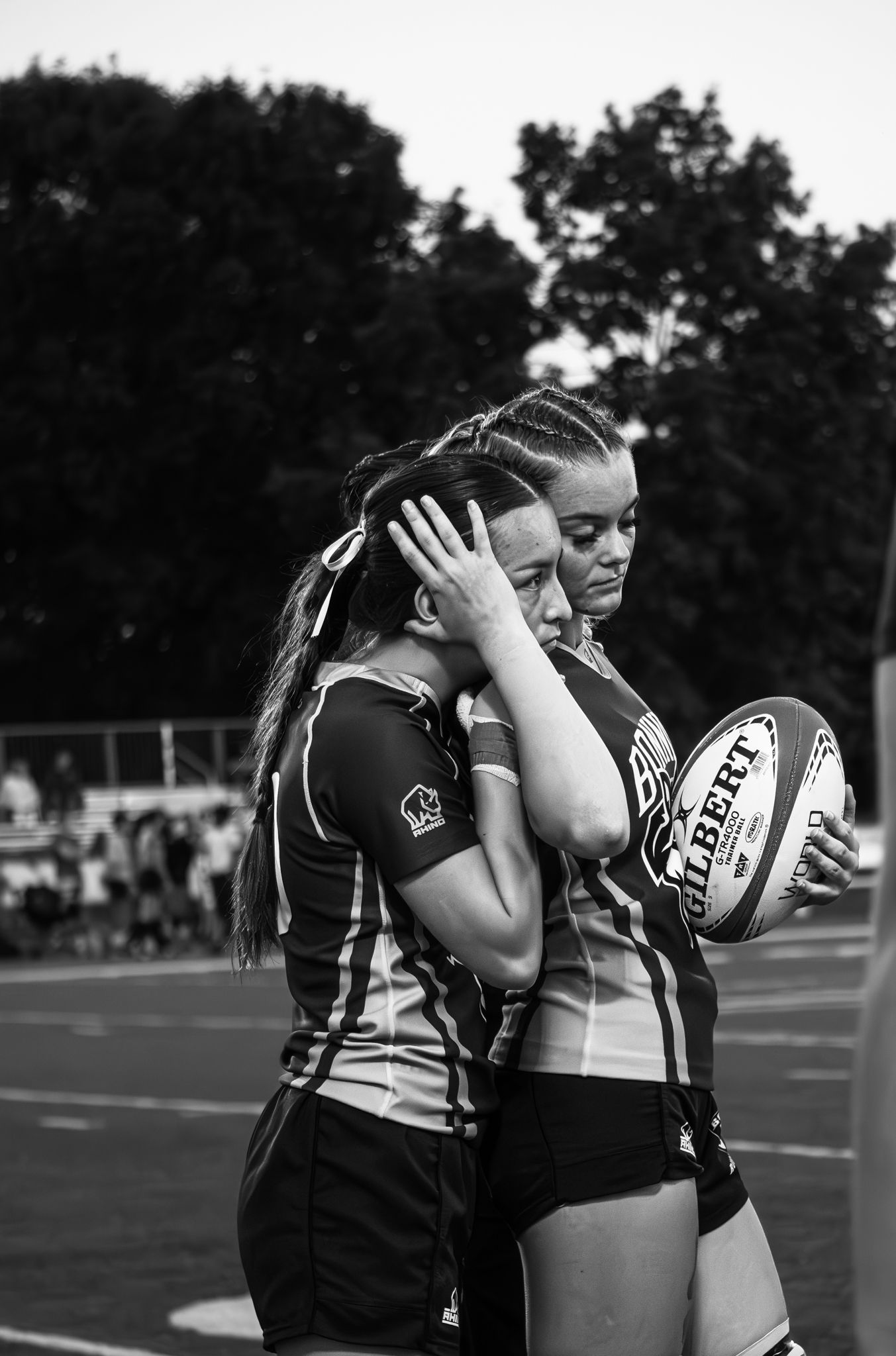 Black and white photo of two rugby players leaning together, one covering her face while the other holds a Gilbert rugby ball.