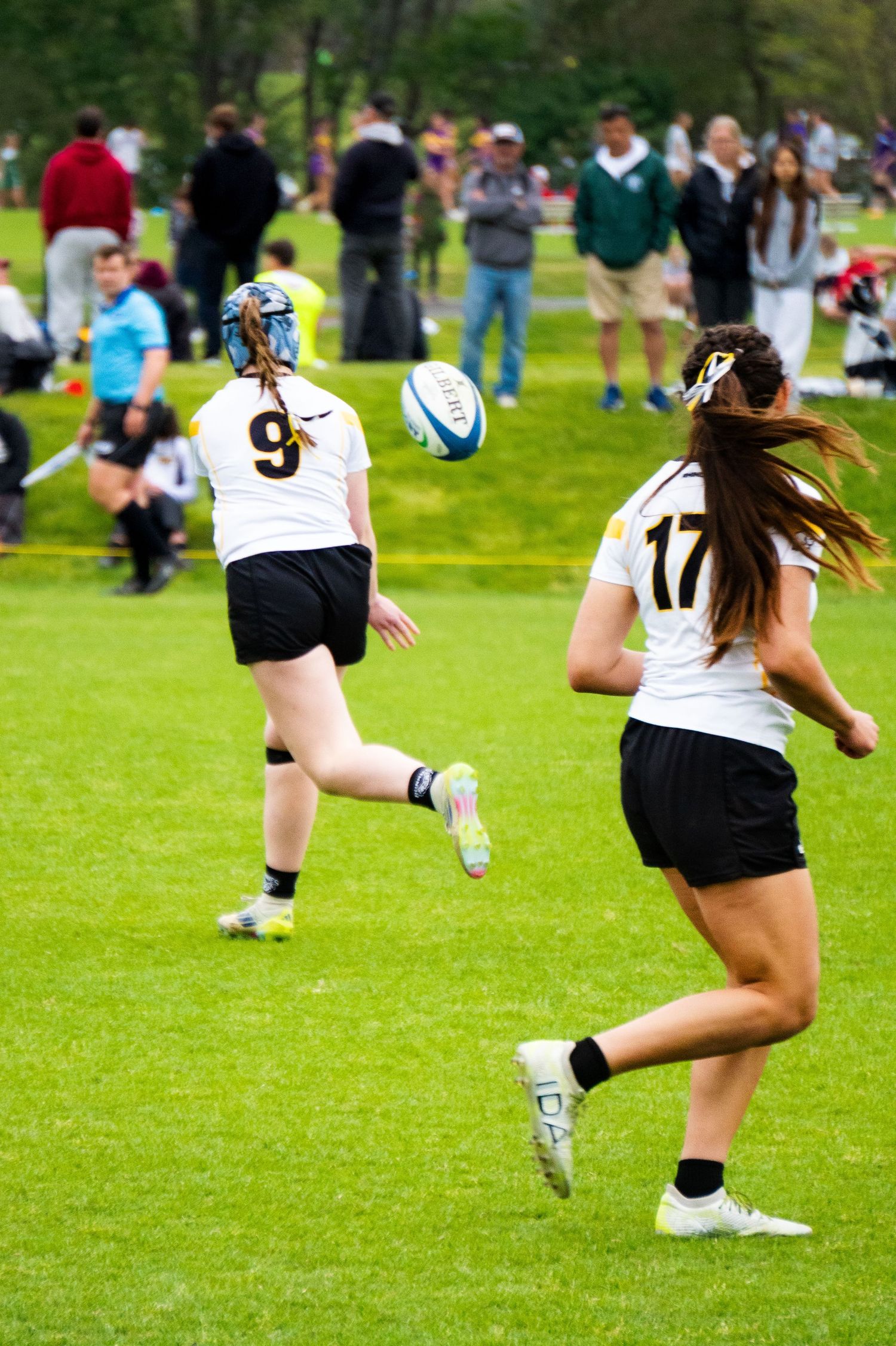 Two women in white jerseys numbered 9 and 17 play rugby on a grass field as a ball sails through the air while spectators watch.