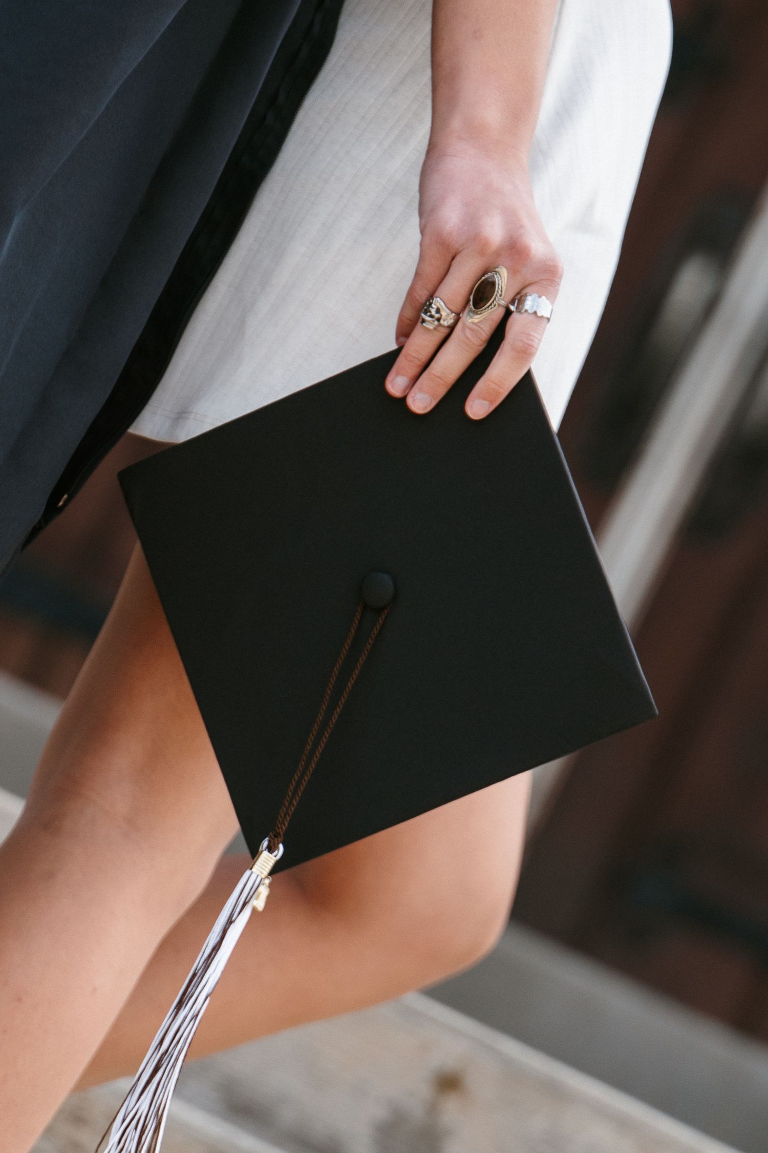 Close-up of a graduate's hand with rings resting on a black graduation cap with a silver tassel against a white dress.