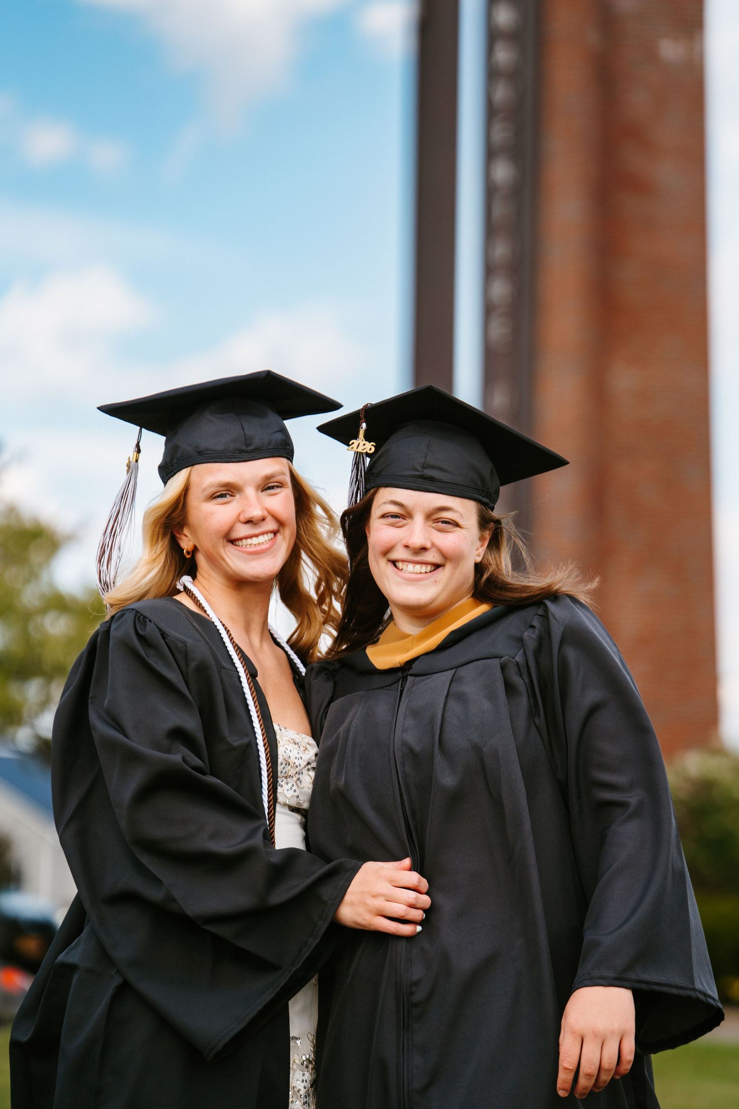 Two smiling graduates in black caps and gowns hug outdoors in front of a brick campus tower under a blue sky.