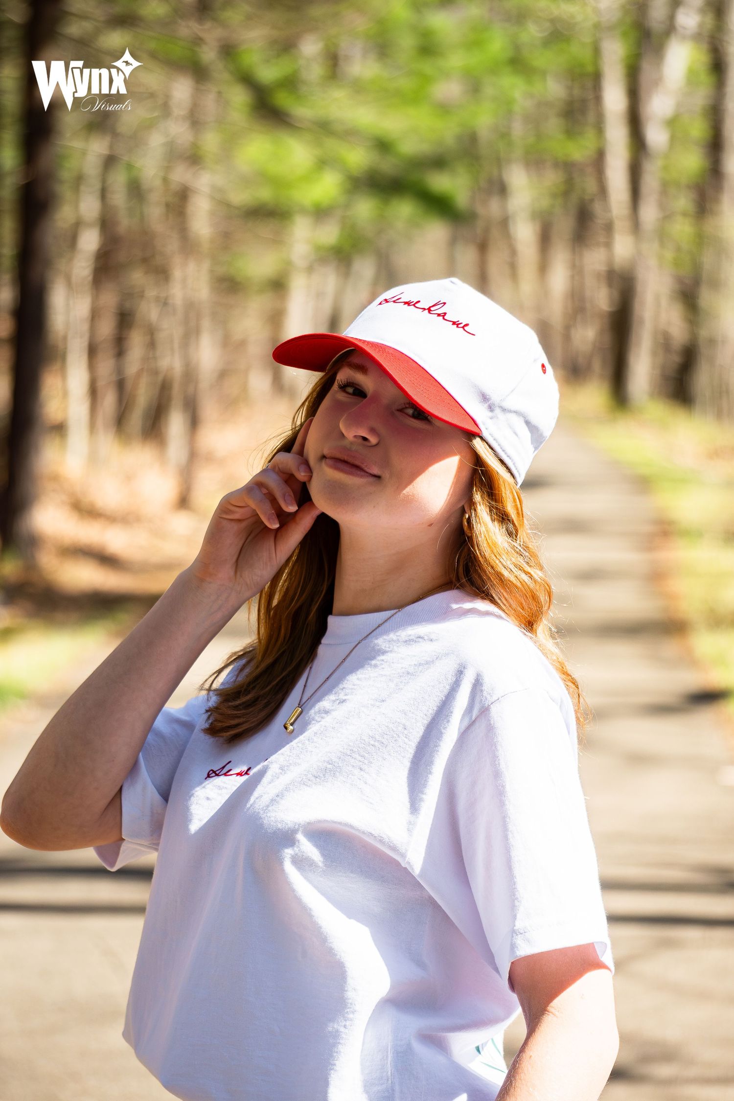 Woman smiling on a tree-lined path, hand at her cheek, wearing a white and red Sewlaw cap and an oversized white tee.