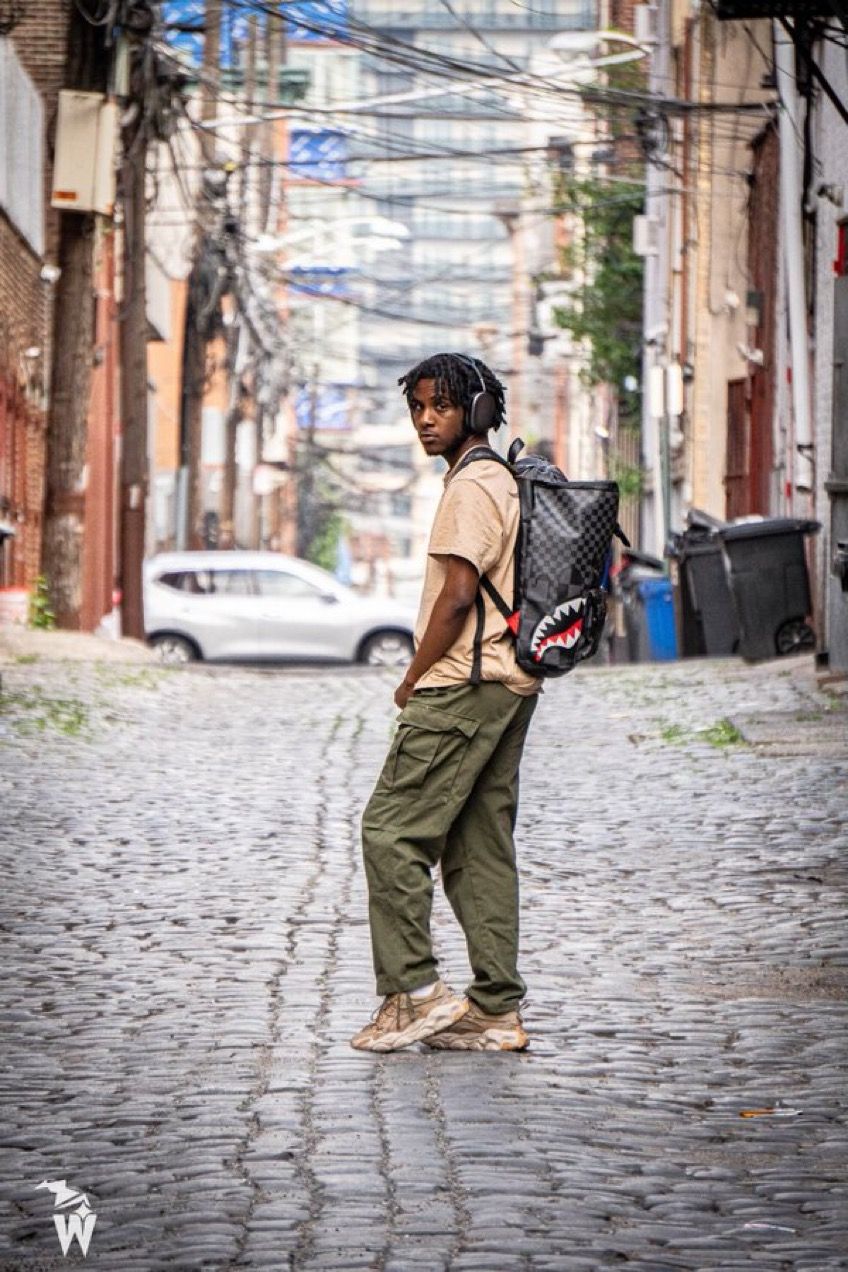Young man in headphones, olive cargo pants and a shark backpack stands on a cobblestone alley with city buildings behind him.