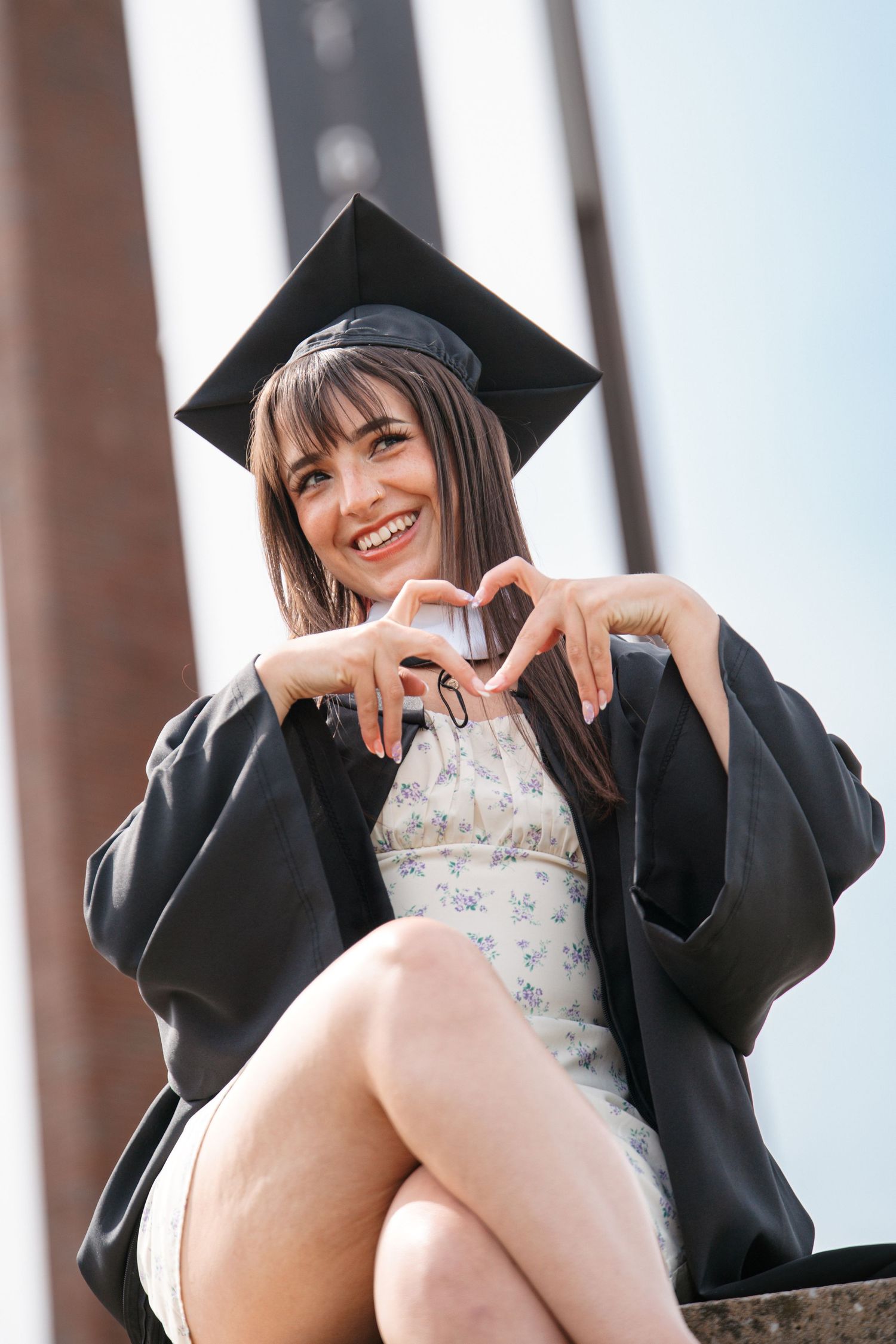 Smiling graduate in a cap and black gown makes a heart shape with her hands while sitting on a stone ledge outdoors.