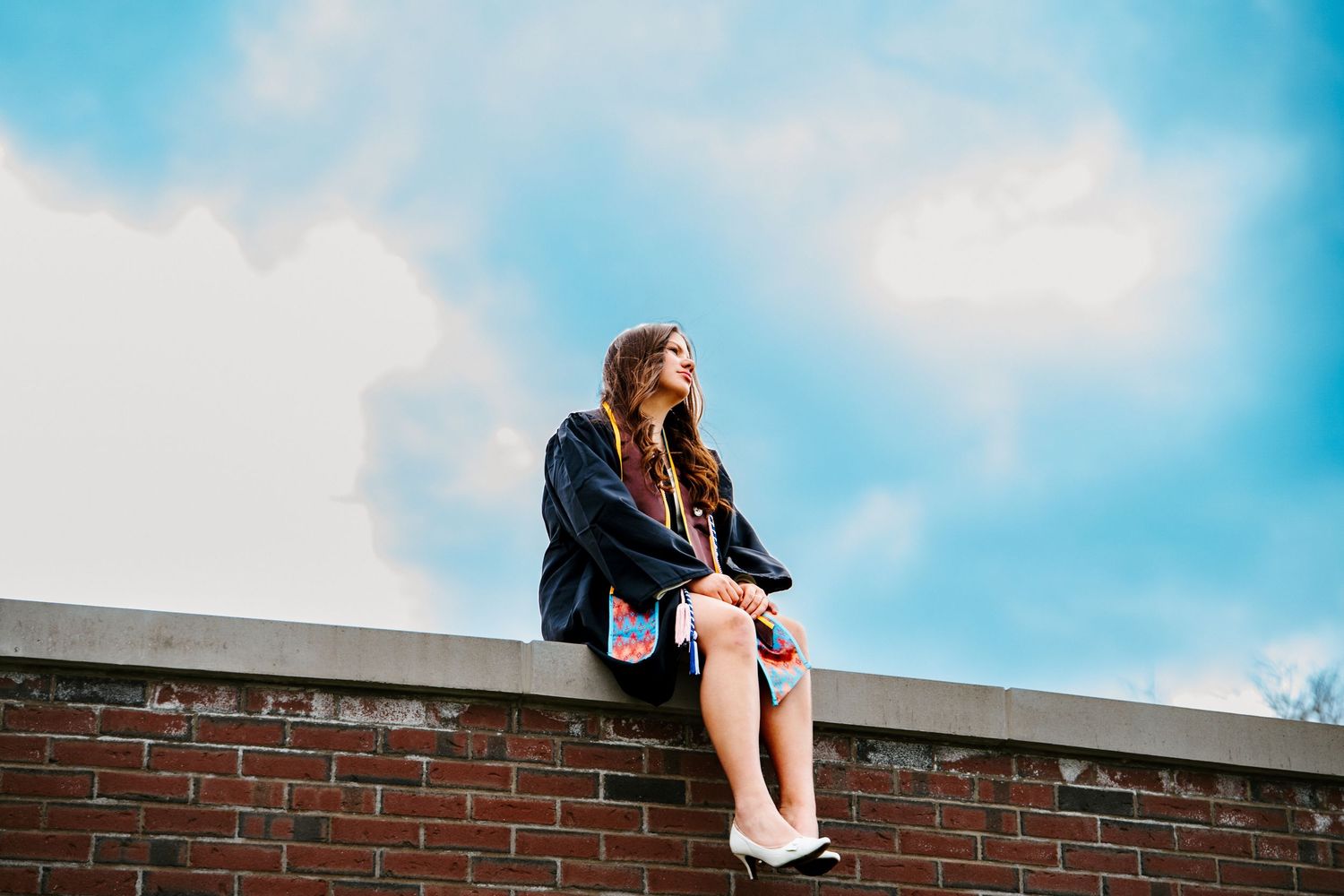 Graduate in a black gown sits on a brick wall and gazes into the distance under a bright blue sky with white clouds.
