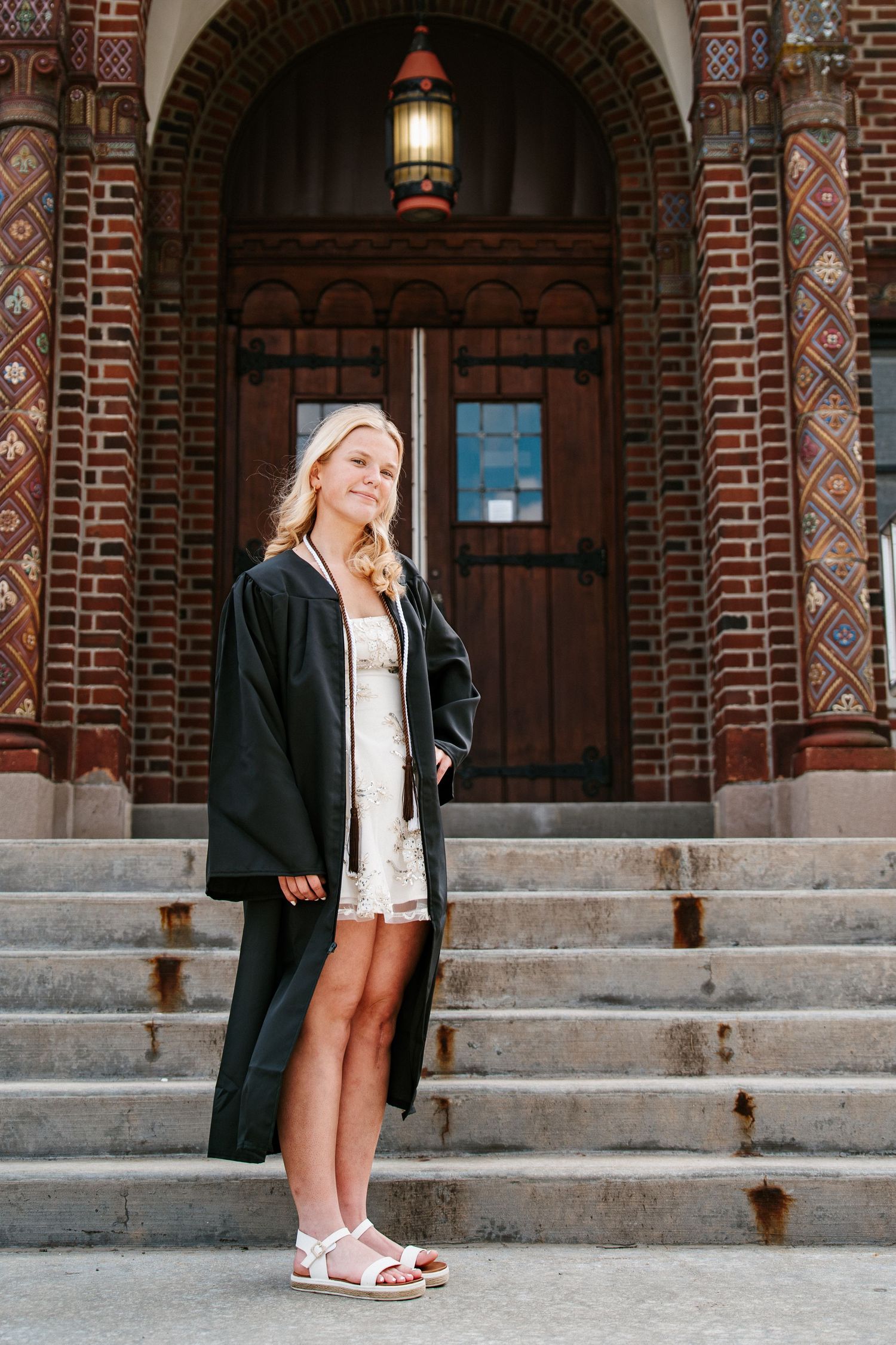 Blonde graduate in a black gown over a white dress and sandals poses on steps in front of an arched brick entrance.