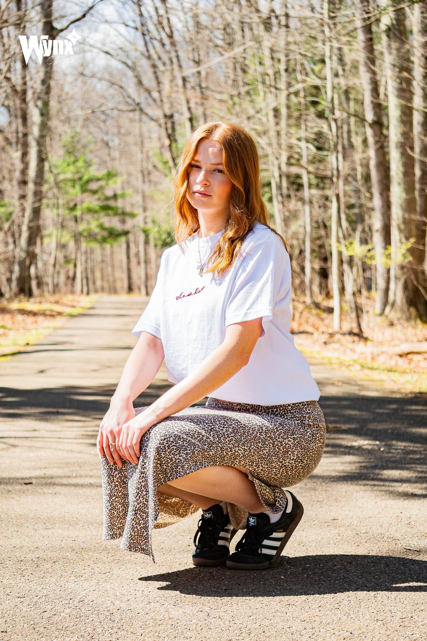 Red-haired woman in a white graphic tee, leopard print skirt and black sneakers crouching on a paved path in a sunlit spring forest.