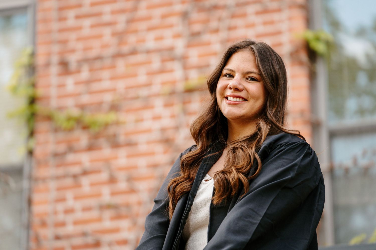 Smiling graduate with long wavy brown hair wears a black gown in front of an ivy-covered red brick building.