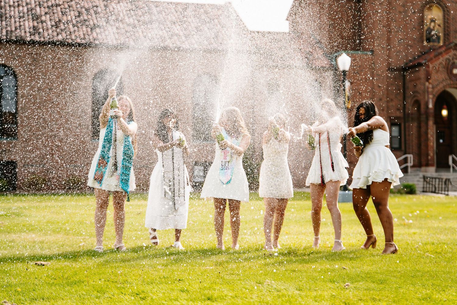 Group of graduates in white dresses celebrate by spraying champagne on a green lawn in front of a historic brick building.