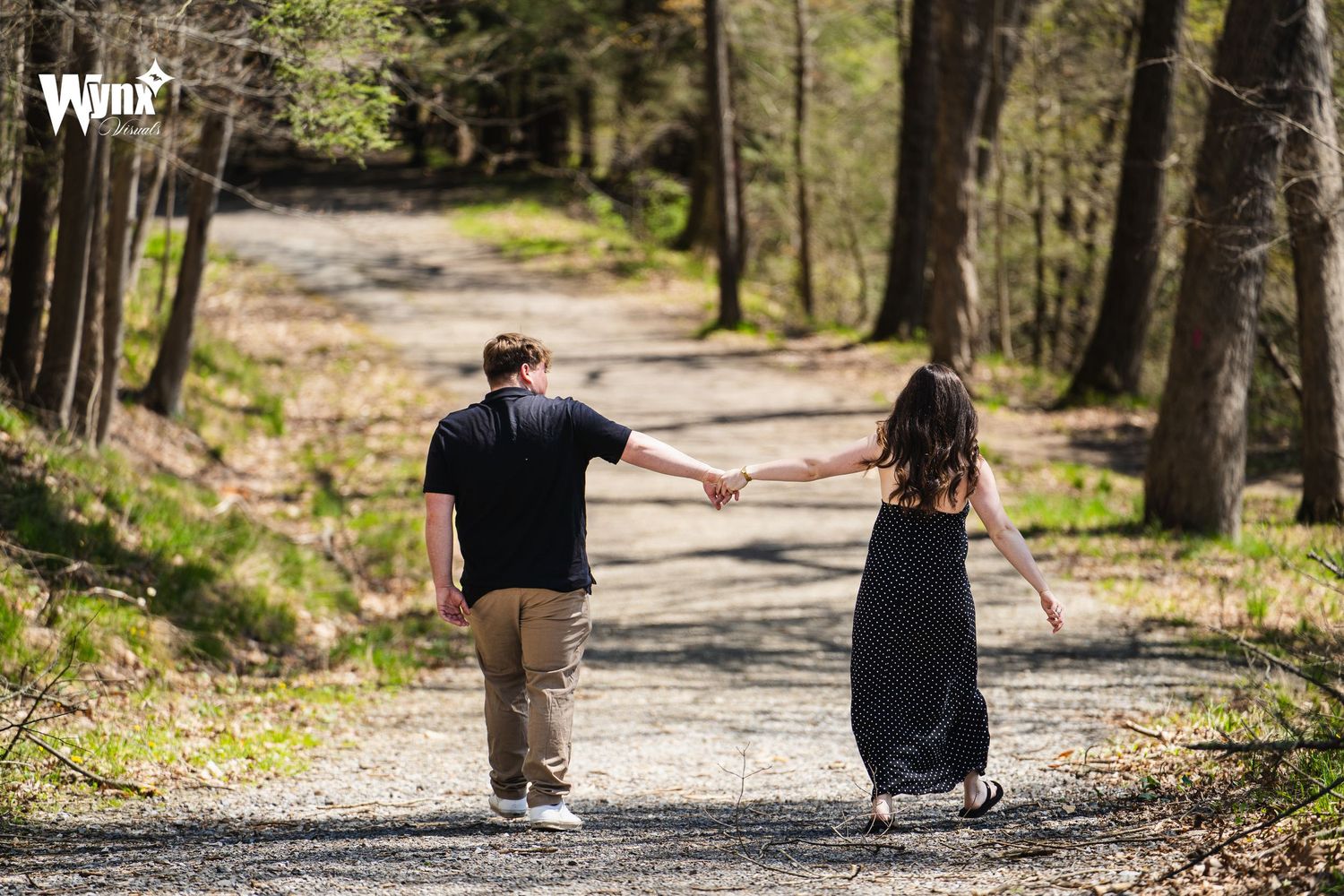 A couple holds hands and walks along a wooded gravel path in sunshine during an outdoor engagement photo session.