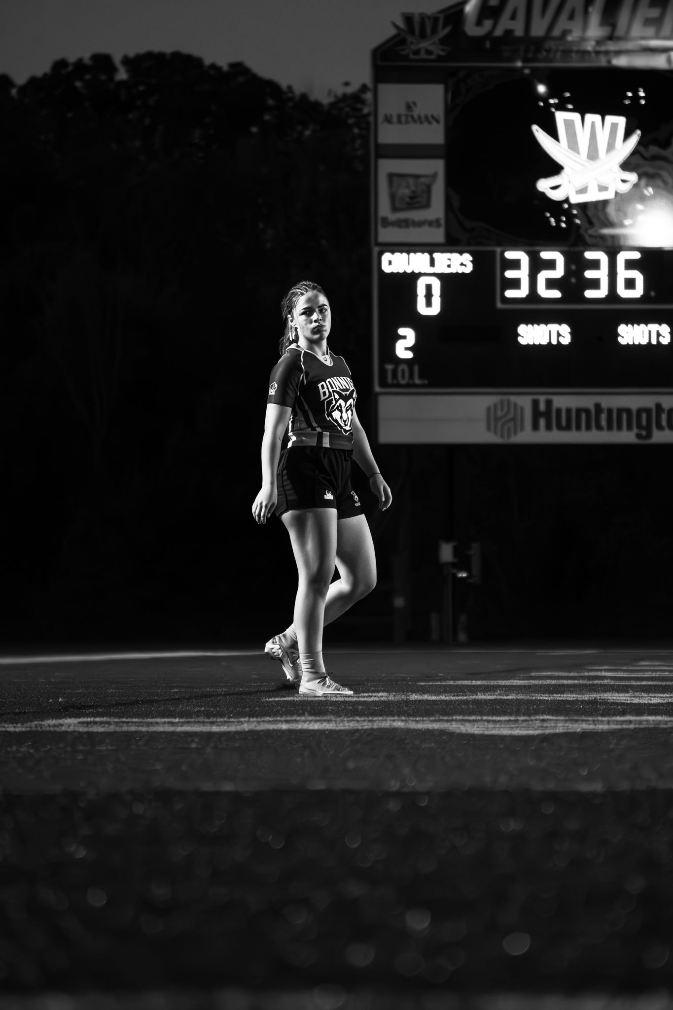 A female rugby player in a dark jersey walks across the field at night in front of a lit Cavaliers scoreboard, in black and white.
