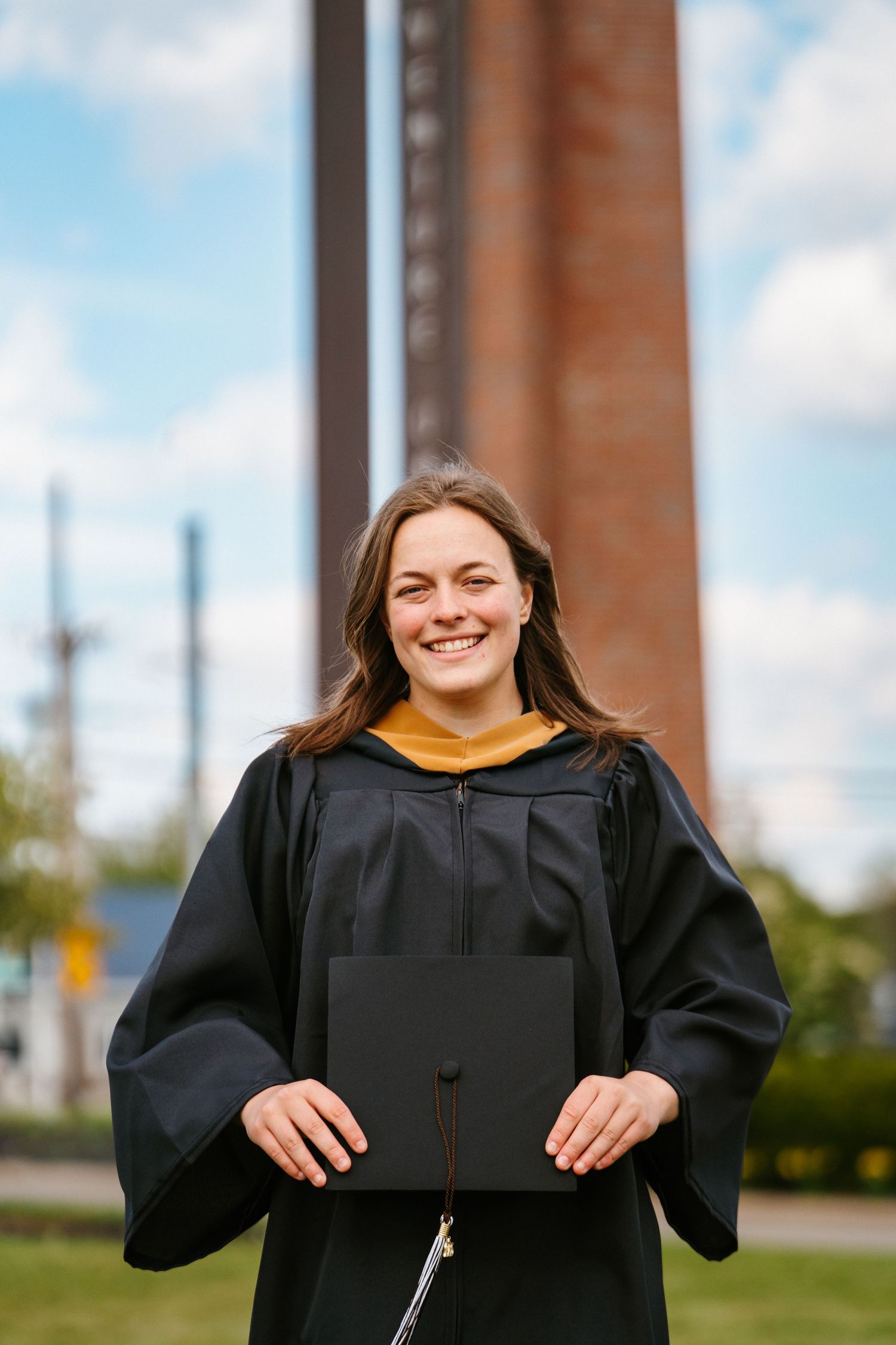 Smiling graduate in a black gown with a gold collar holds her mortarboard cap in front of a campus brick tower.