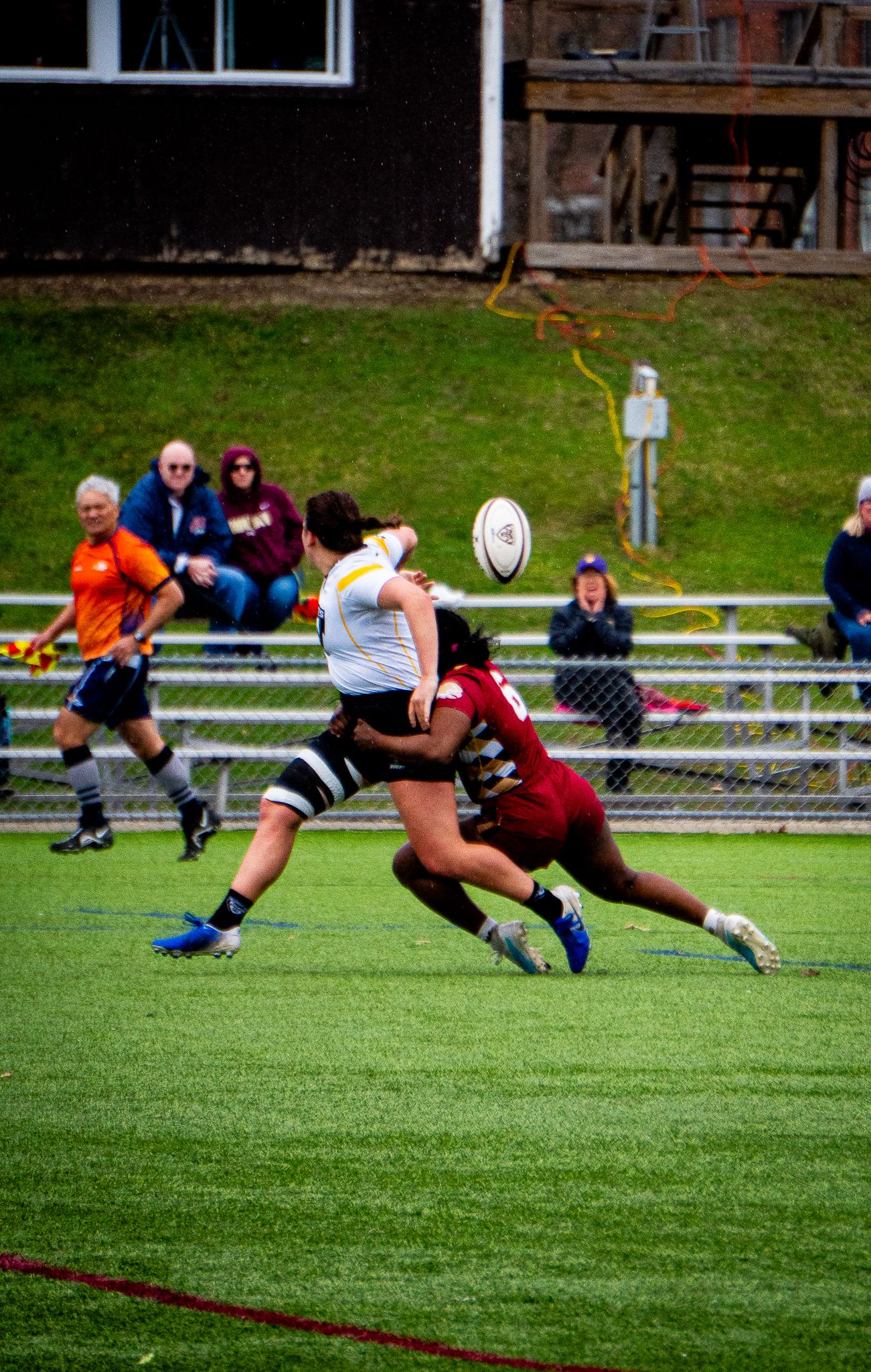 A women's rugby player in a white jersey is tackled by a defender in maroon as the ball flies overhead and a referee and fans watch.