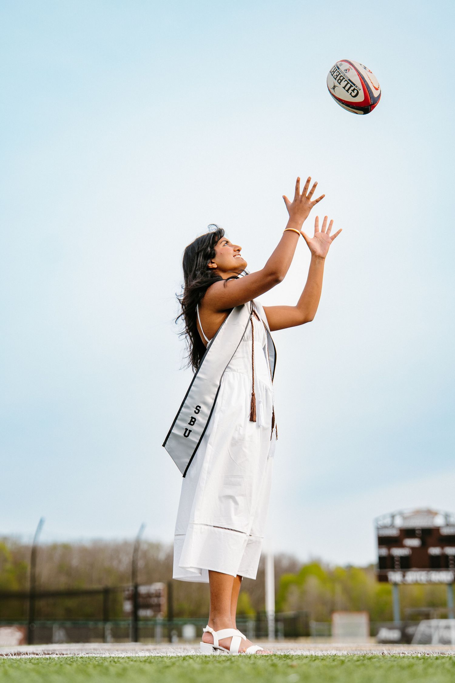 A graduate in a white dress and stole reaches up to catch a rugby ball against a clear sky on a sports field.
