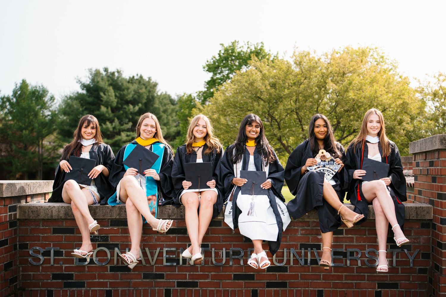 Six female graduates in black caps and gowns sit in a row on a brick wall, smiling and holding diplomas among autumn trees.