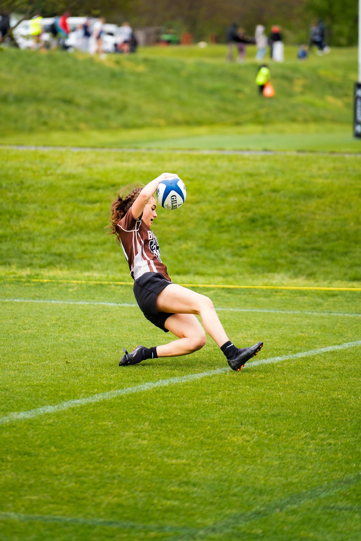 A women's rugby player in a brown jersey slides along the grass and stretches her arm to ground the ball for a try.