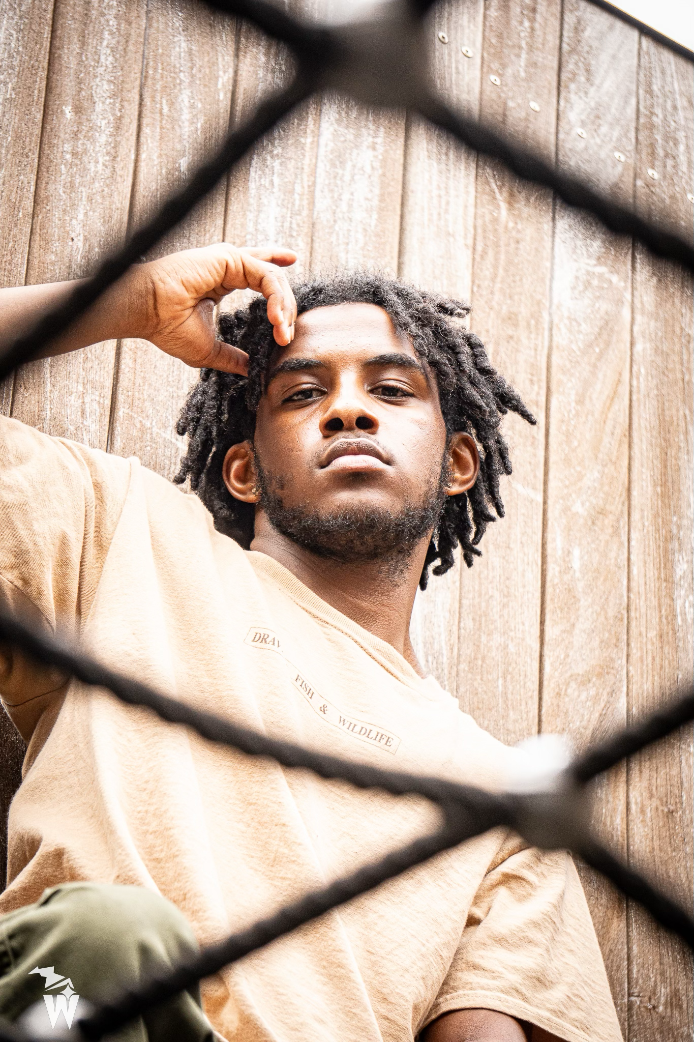Young man with short locs and a beard poses in a tan t-shirt, hand raised to his head, photographed through a chain-link fence.