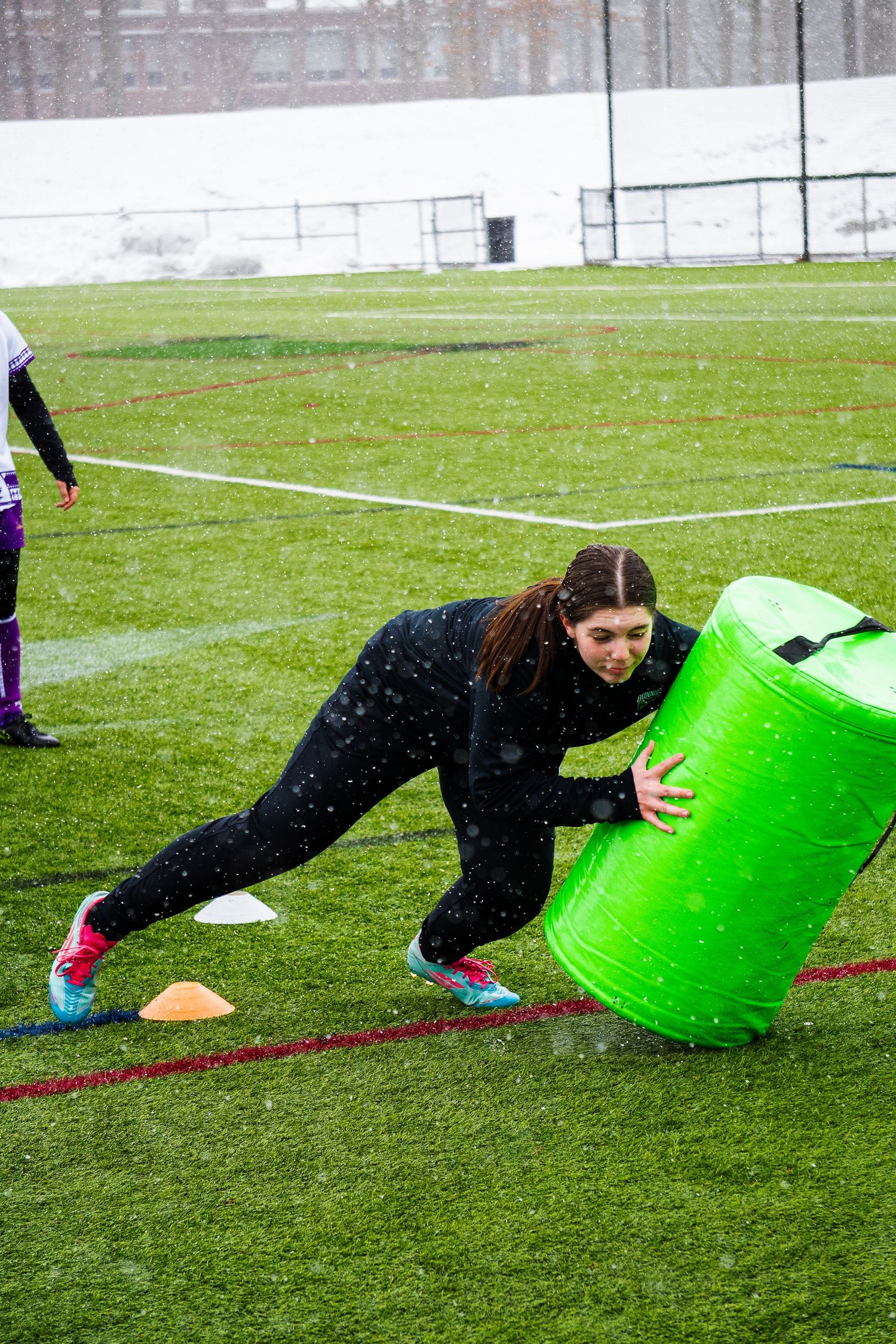 A women's rugby player in black leans into a green tackle bag during a snowy training drill on a turf field.
