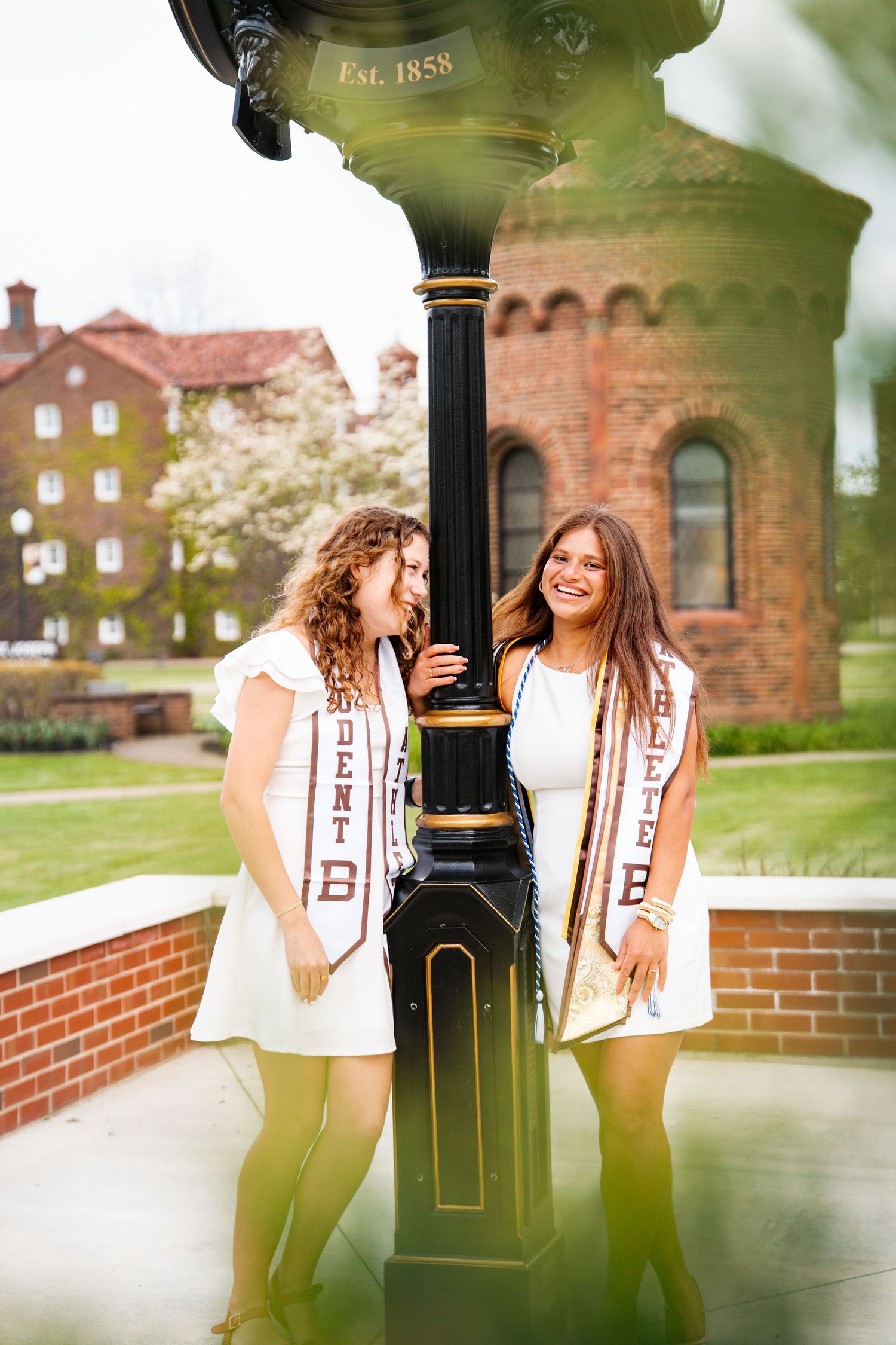Two smiling graduates in white dresses and maroon sashes pose beside a black lamppost near brick campus buildings.
