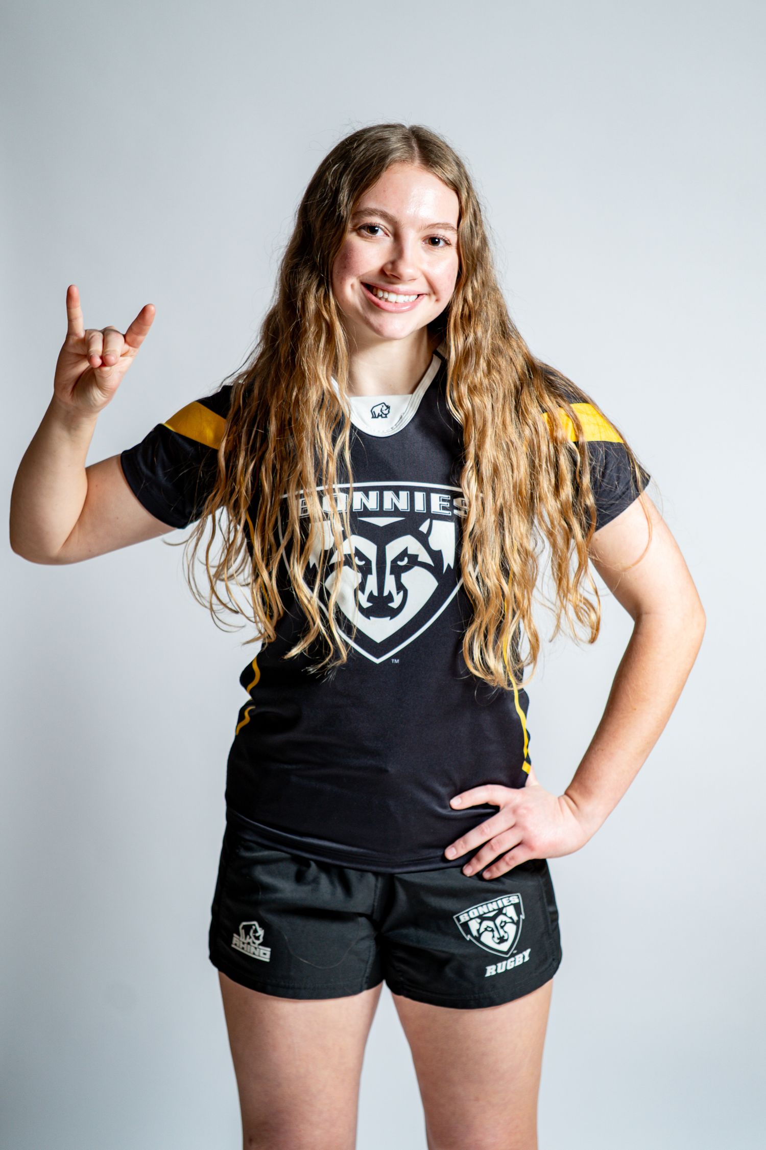 Smiling rugby player with long wavy hair in a black Bonnies jersey and shorts makes a rock-on hand gesture against a white backdrop.