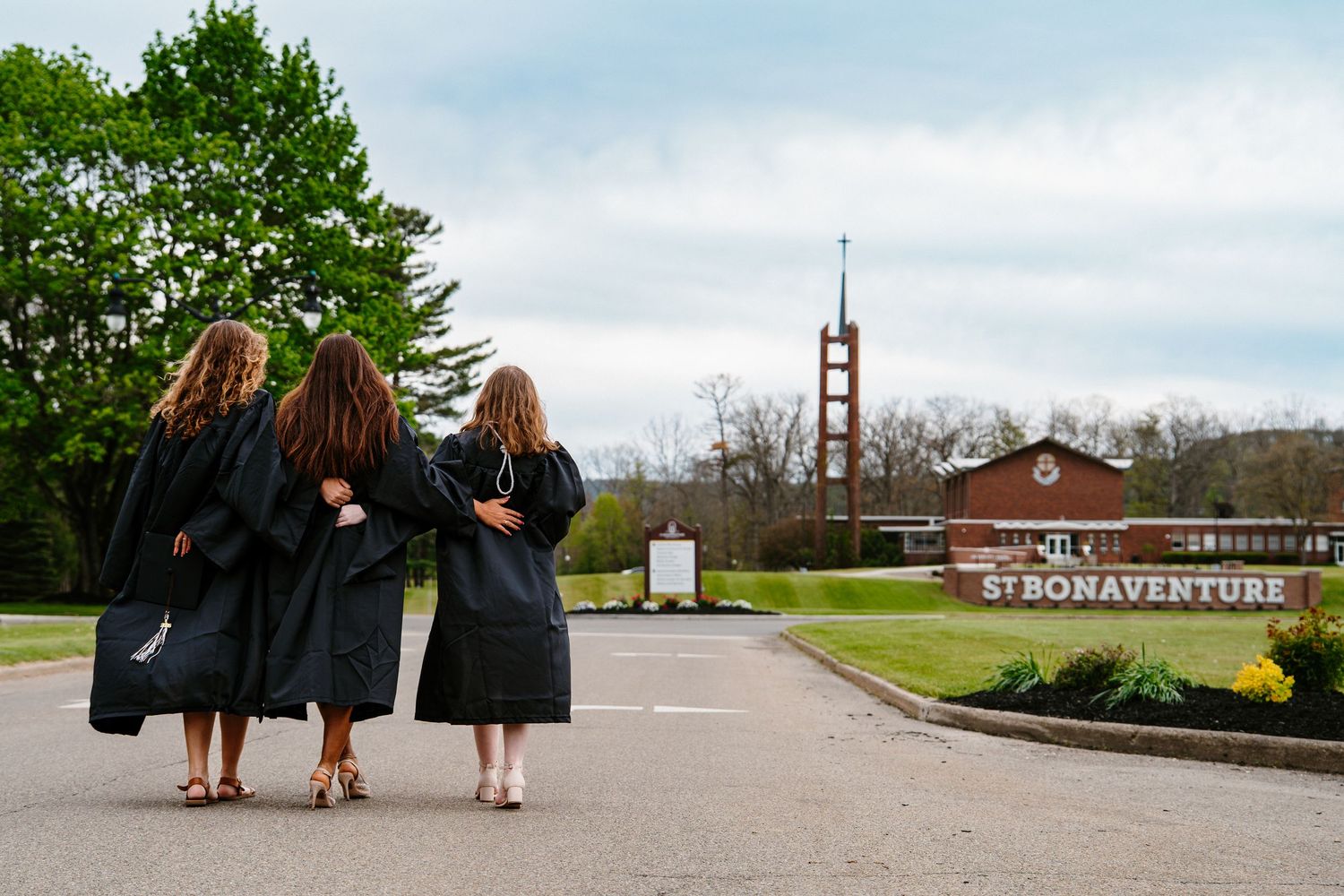 Three graduates in black gowns link arms and walk toward the St. Bonaventure University sign and bell tower on a cloudy day.