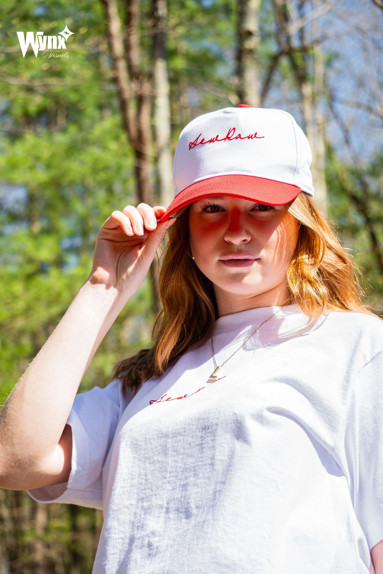 Woman holding the brim of her white and red Sewlaw cap while wearing a gold pendant necklace and white tee in the woods.