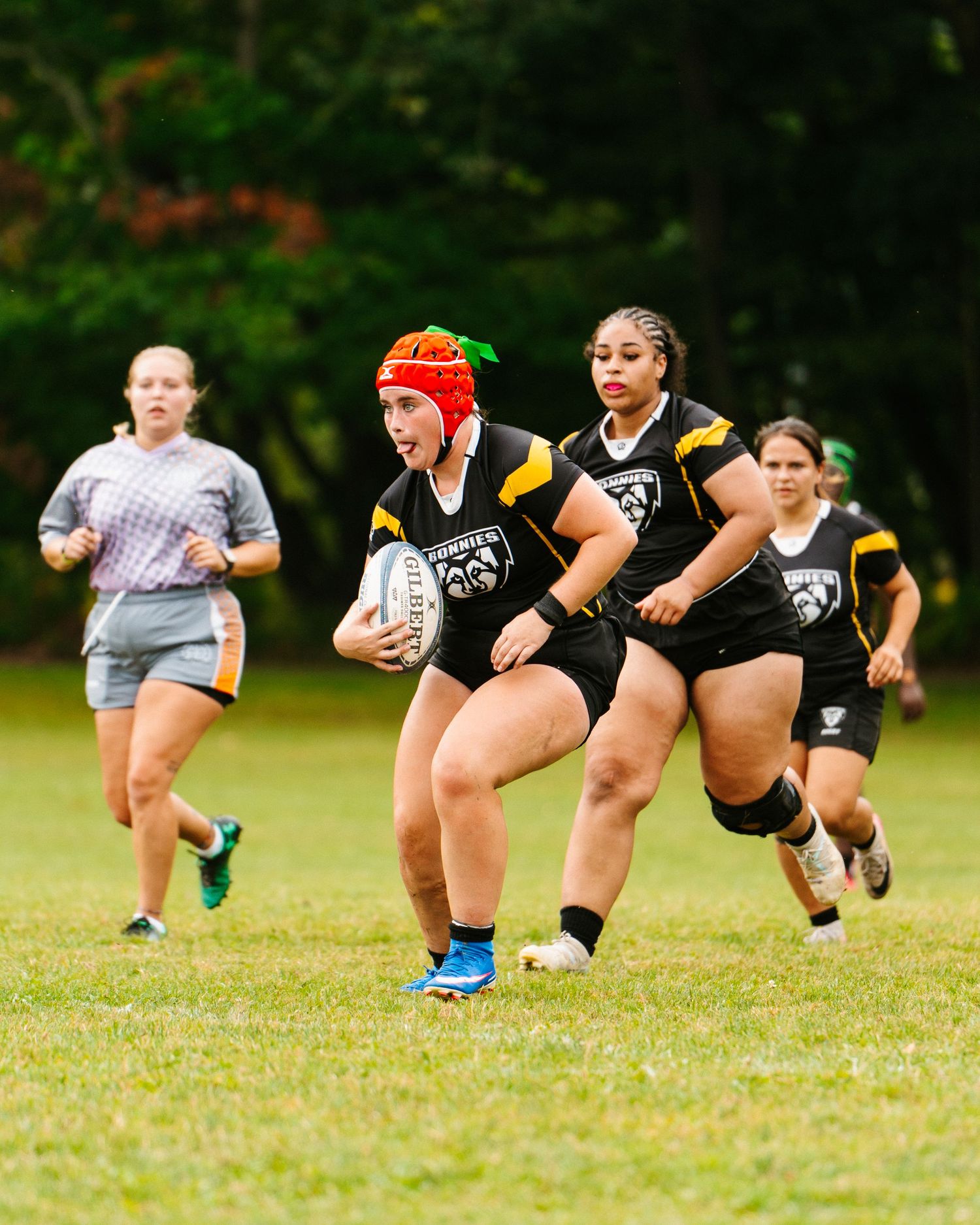 Women's rugby player in a black and gold uniform and orange headgear carries the ball as teammates run alongside her.