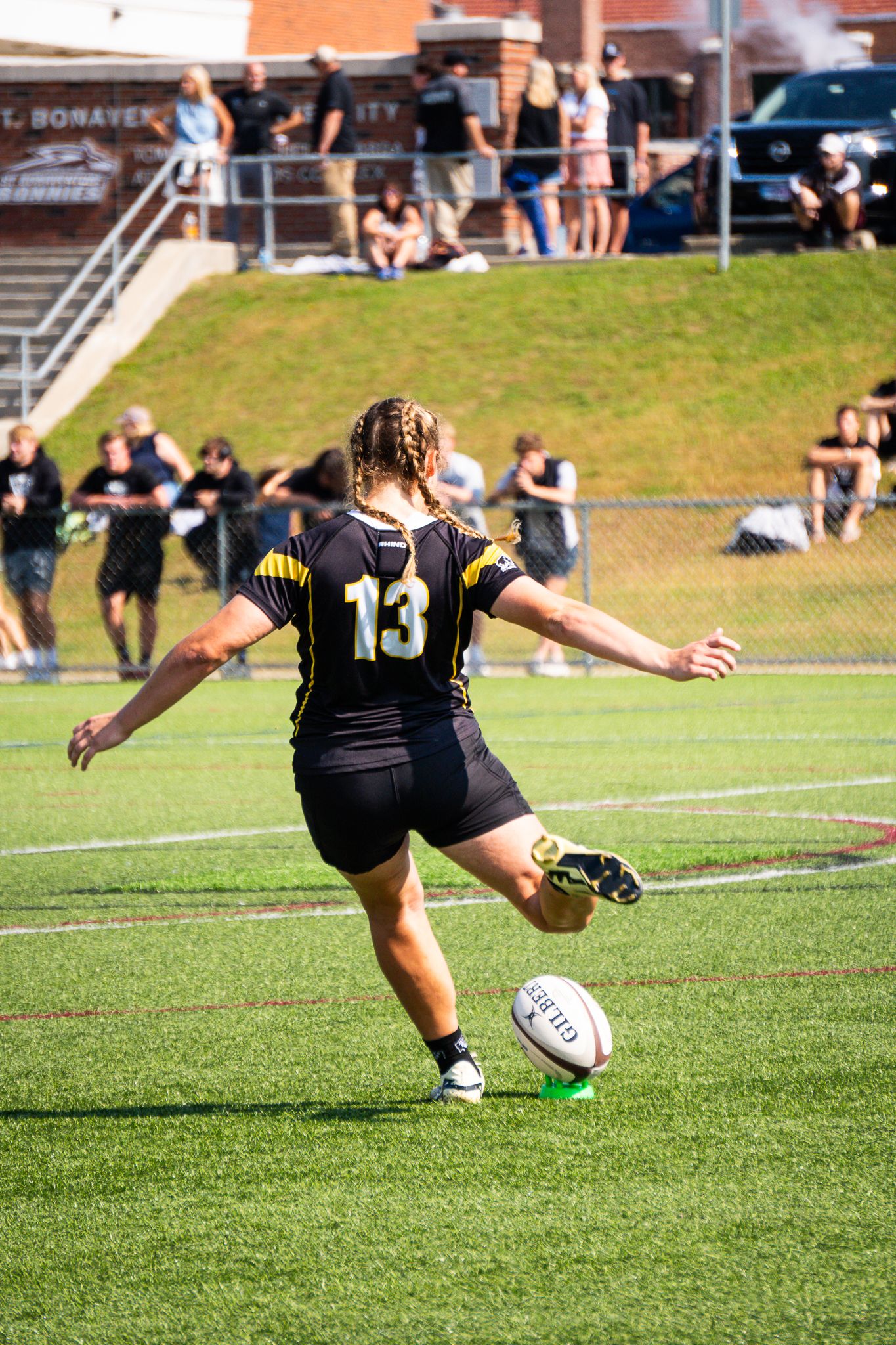 A women's rugby player wearing number 13 in a black jersey lines up to kick the ball from a tee as spectators watch from the hill.