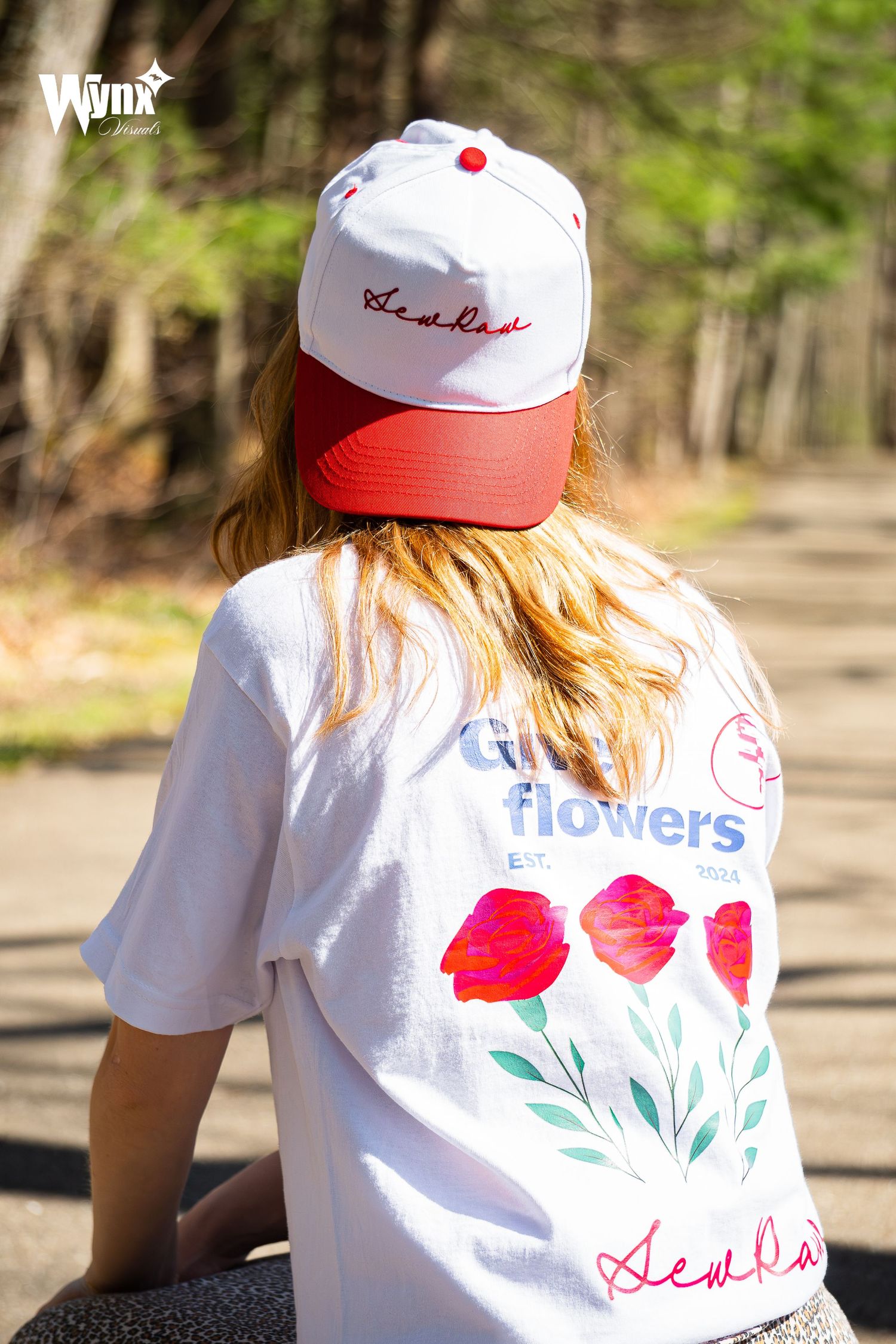 Woman seen from behind wearing a white and red Sewlaw cap and a white Girl Flowers tee with red tulip graphics on a wooded path.