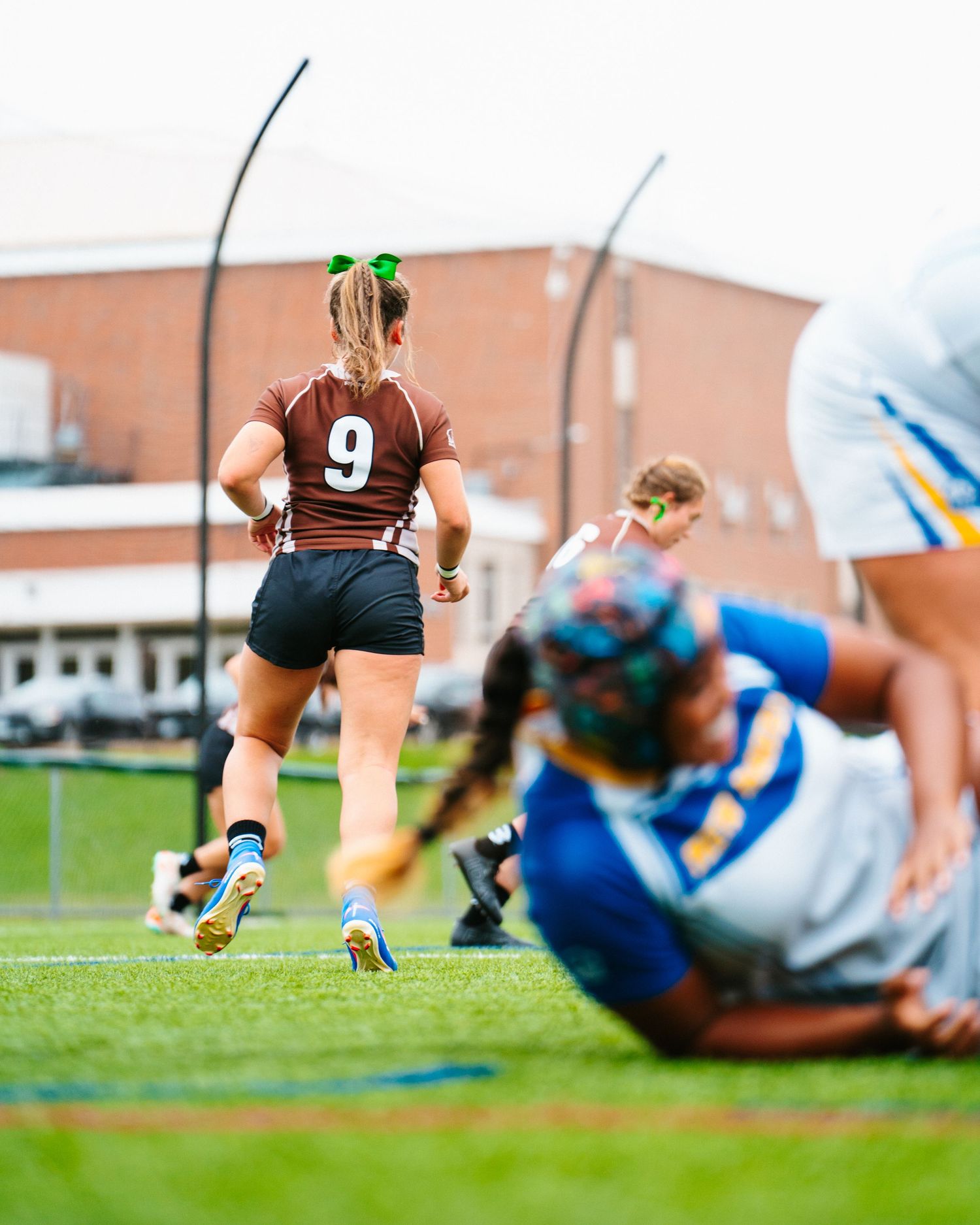 Female rugby player in a brown number 9 jersey runs across the field while opposing players in blue tackle on the grass.