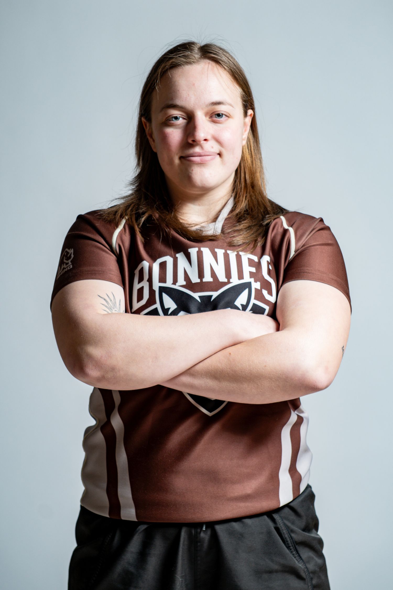 Rugby player with long hair stands with crossed arms in a brown Bonnies jersey featuring a wolf logo against a gray studio background.