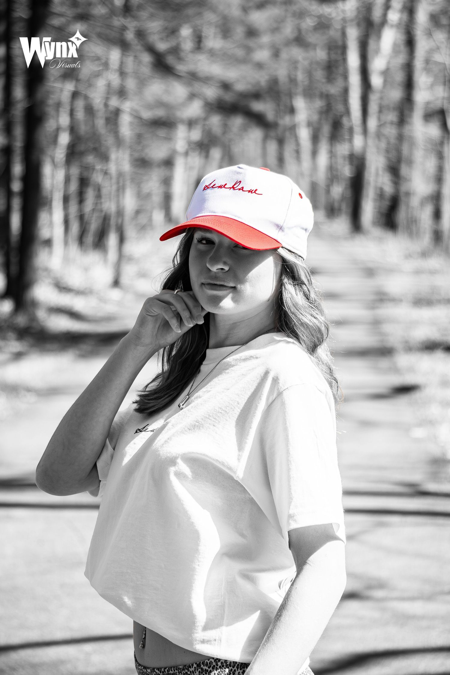 Monochrome forest portrait of a woman in a cropped white tee and Sewlaw cap with red brim and embroidery as the only color.