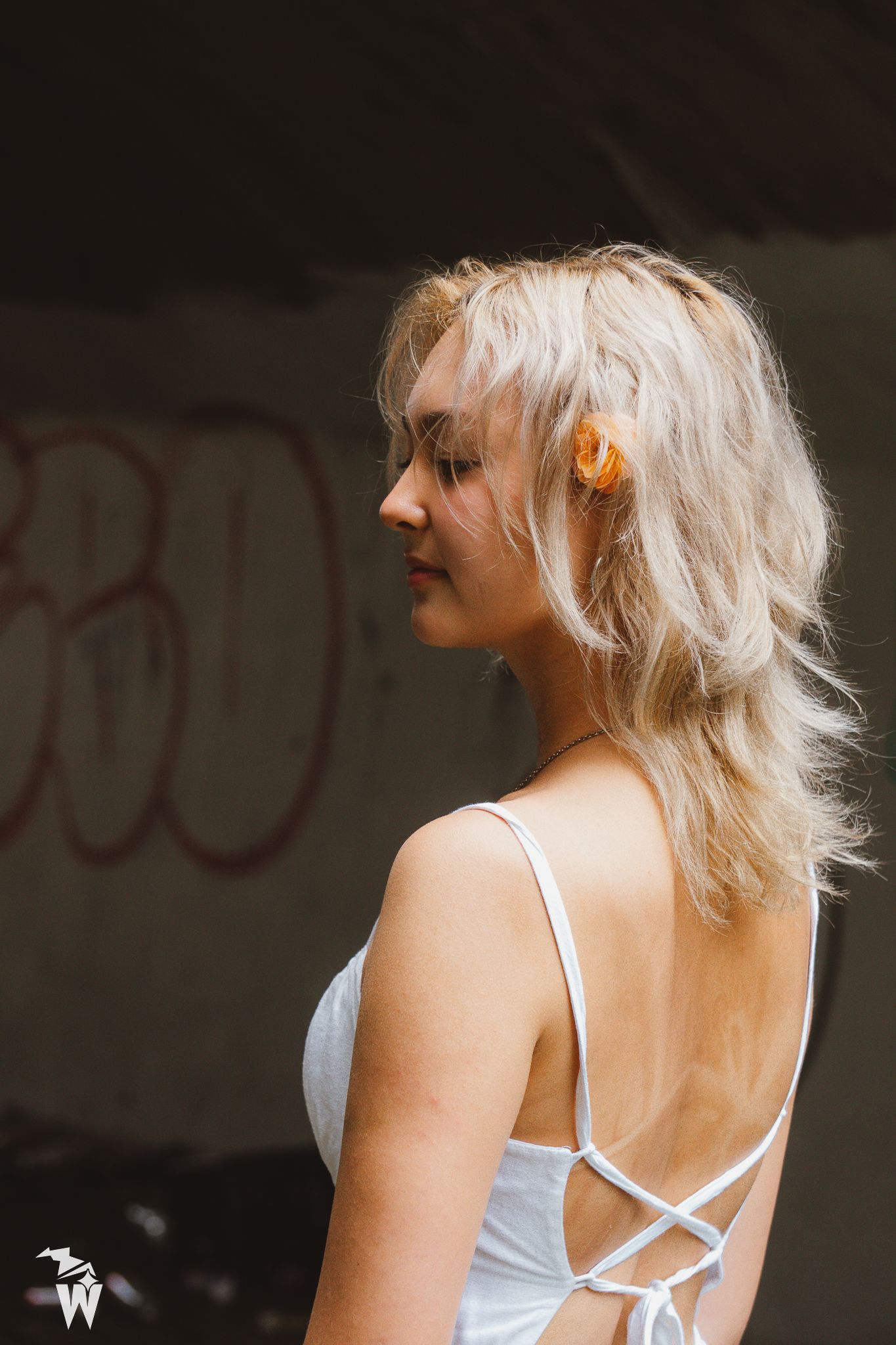 Profile view of a blonde woman in a white backless dress with a flower in her hair, standing before a graffiti wall.