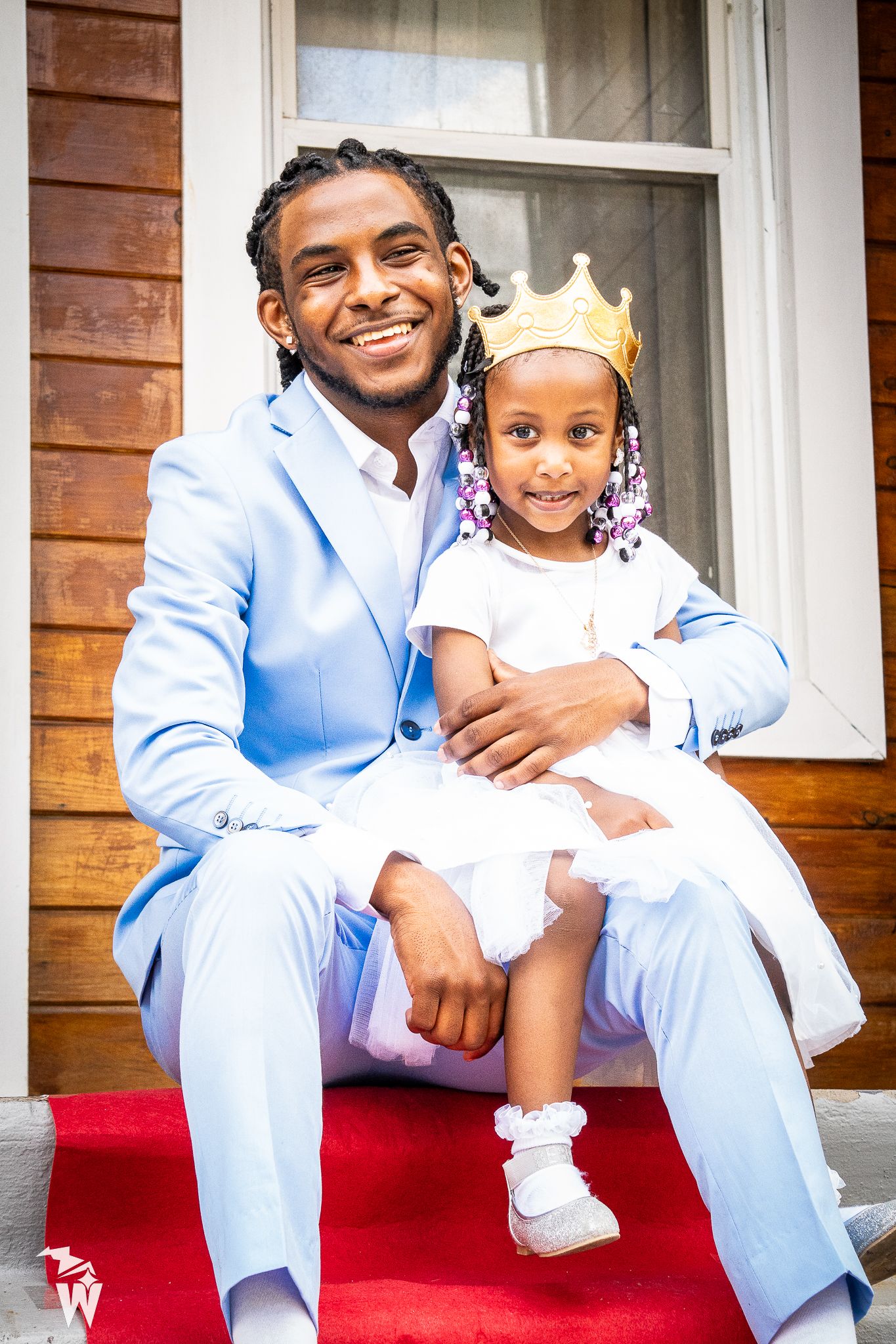 Smiling man in a light blue suit holds a little girl wearing a gold crown on red-carpeted steps outside a wooden house.