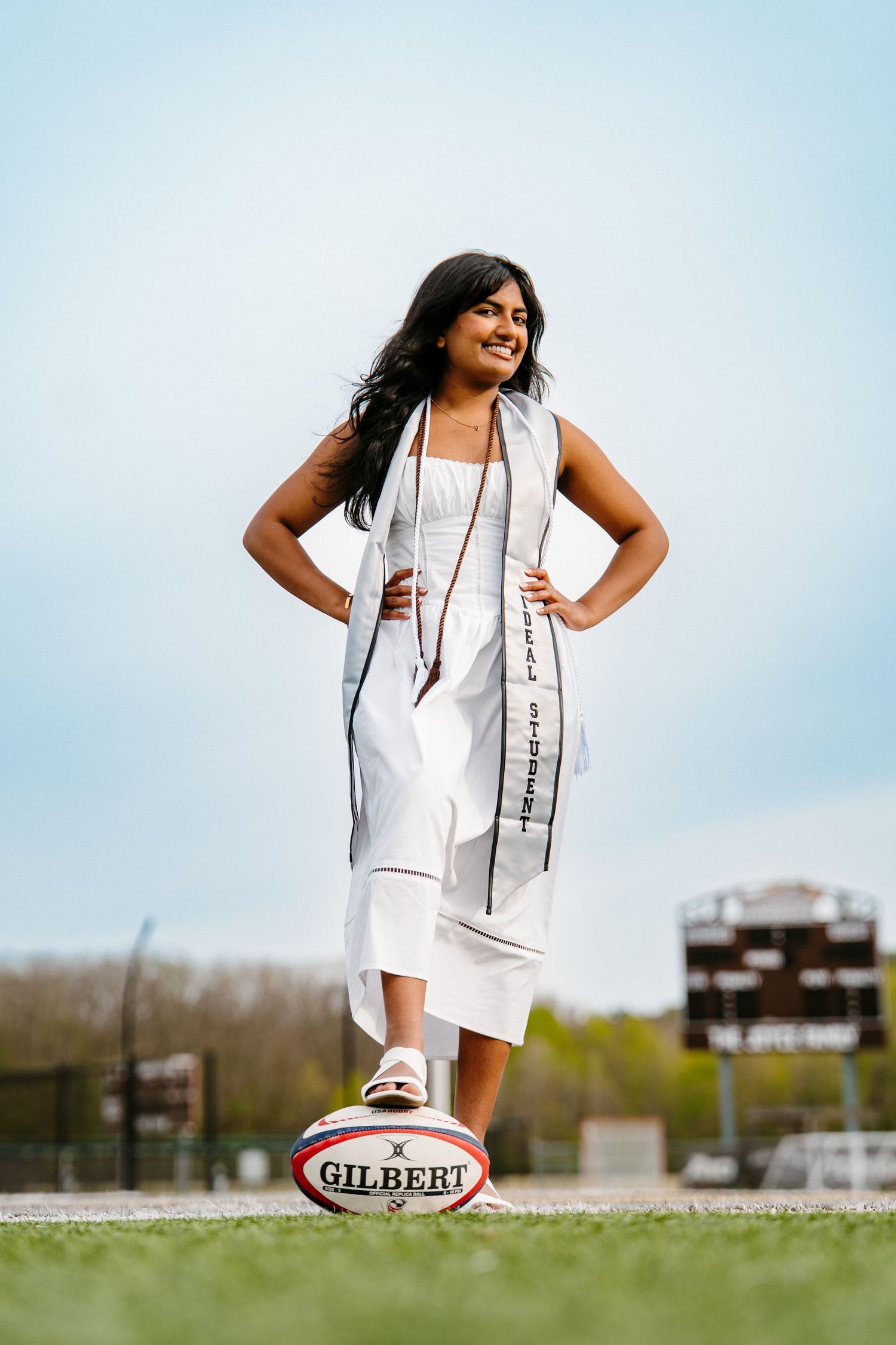 A graduate in a white dress and Student Athlete stole poses with hands on hips and one foot on a Gilbert rugby ball on a field.