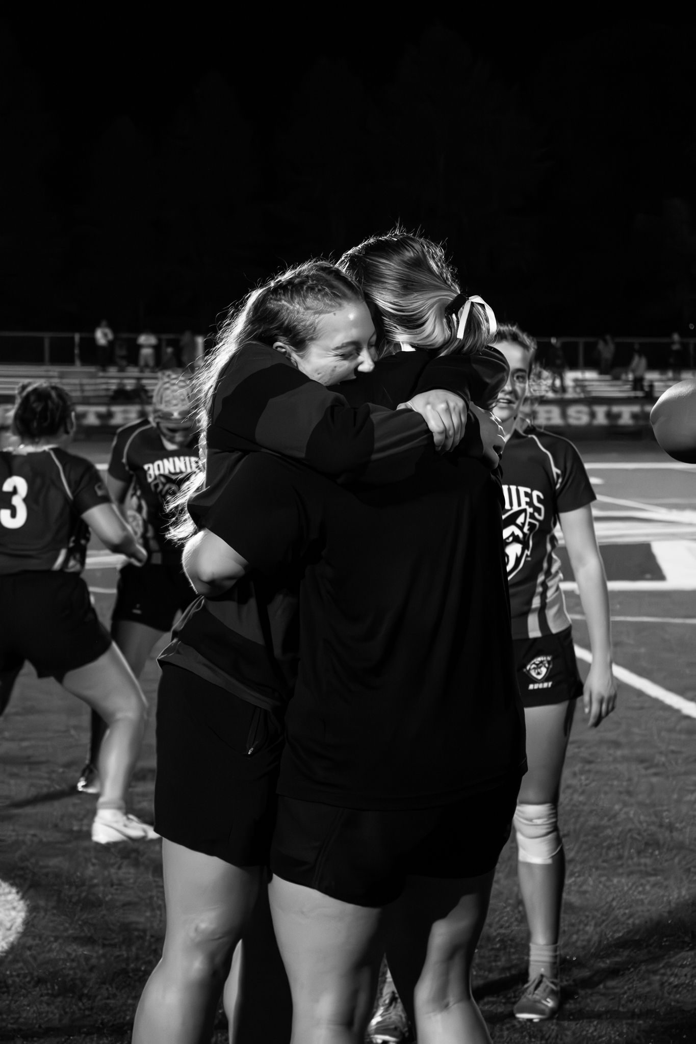 Two rugby players share an emotional hug on the field at night while teammates stand nearby, captured in black and white.