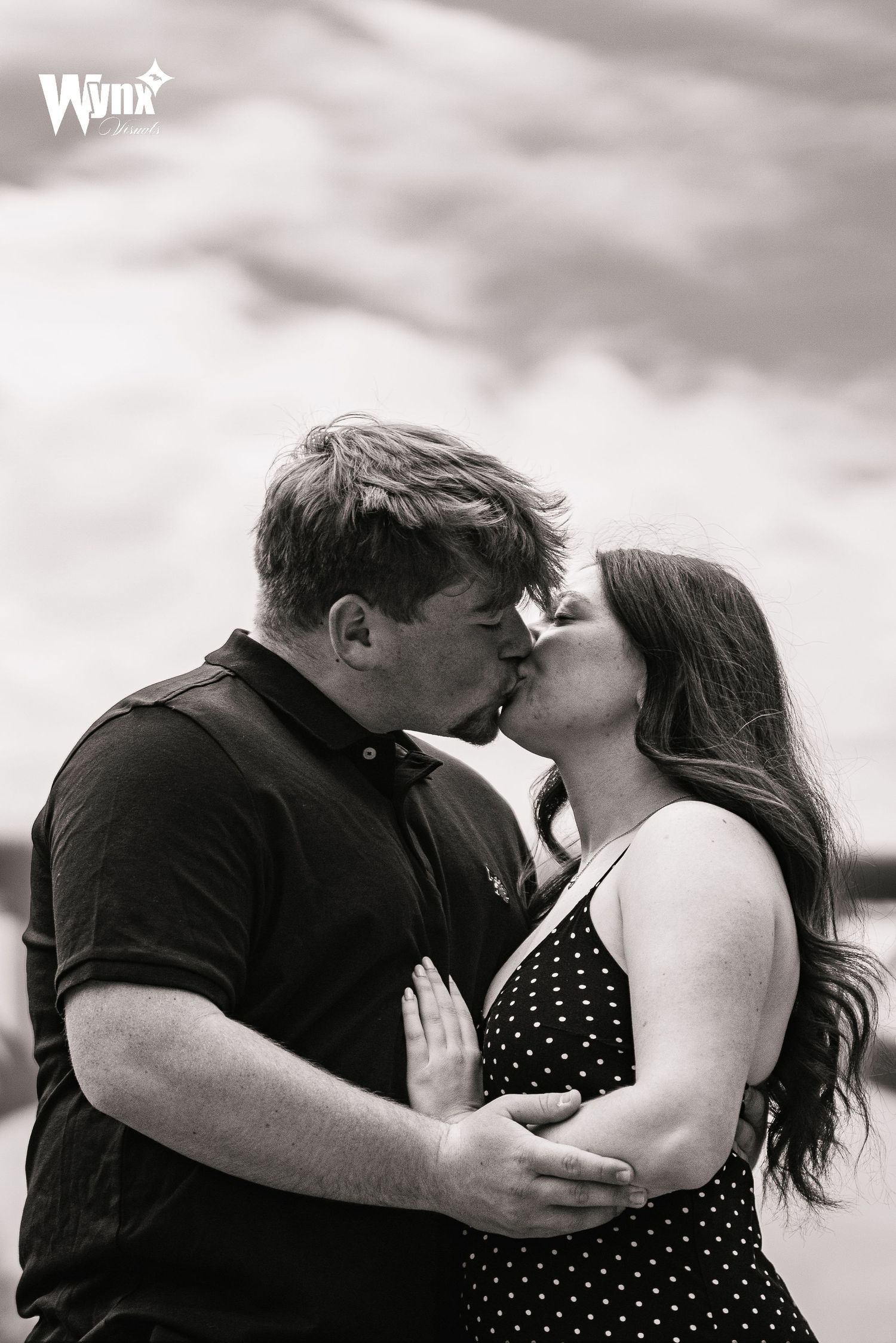 Black and white photo of a couple sharing a kiss outdoors under a cloudy sky, the man in a polo and the woman in a polka dot dress.