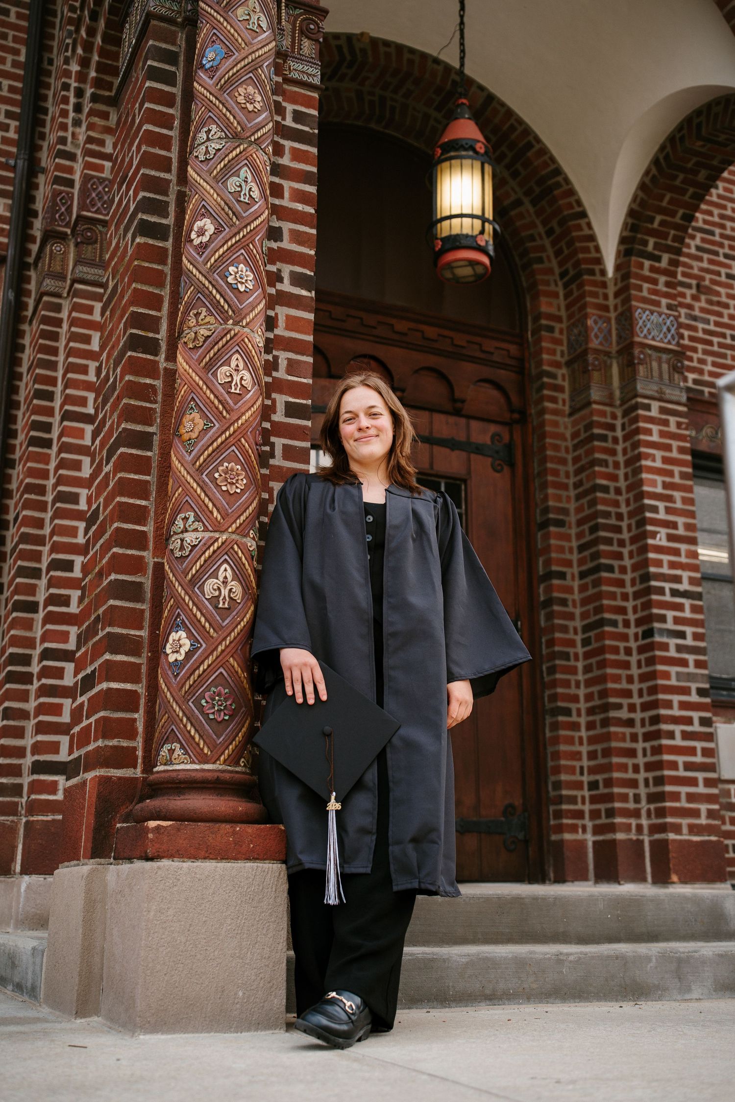 Graduate in a black gown holds her cap and leans on a decorative brick column beside an arched wooden door and hanging lantern.