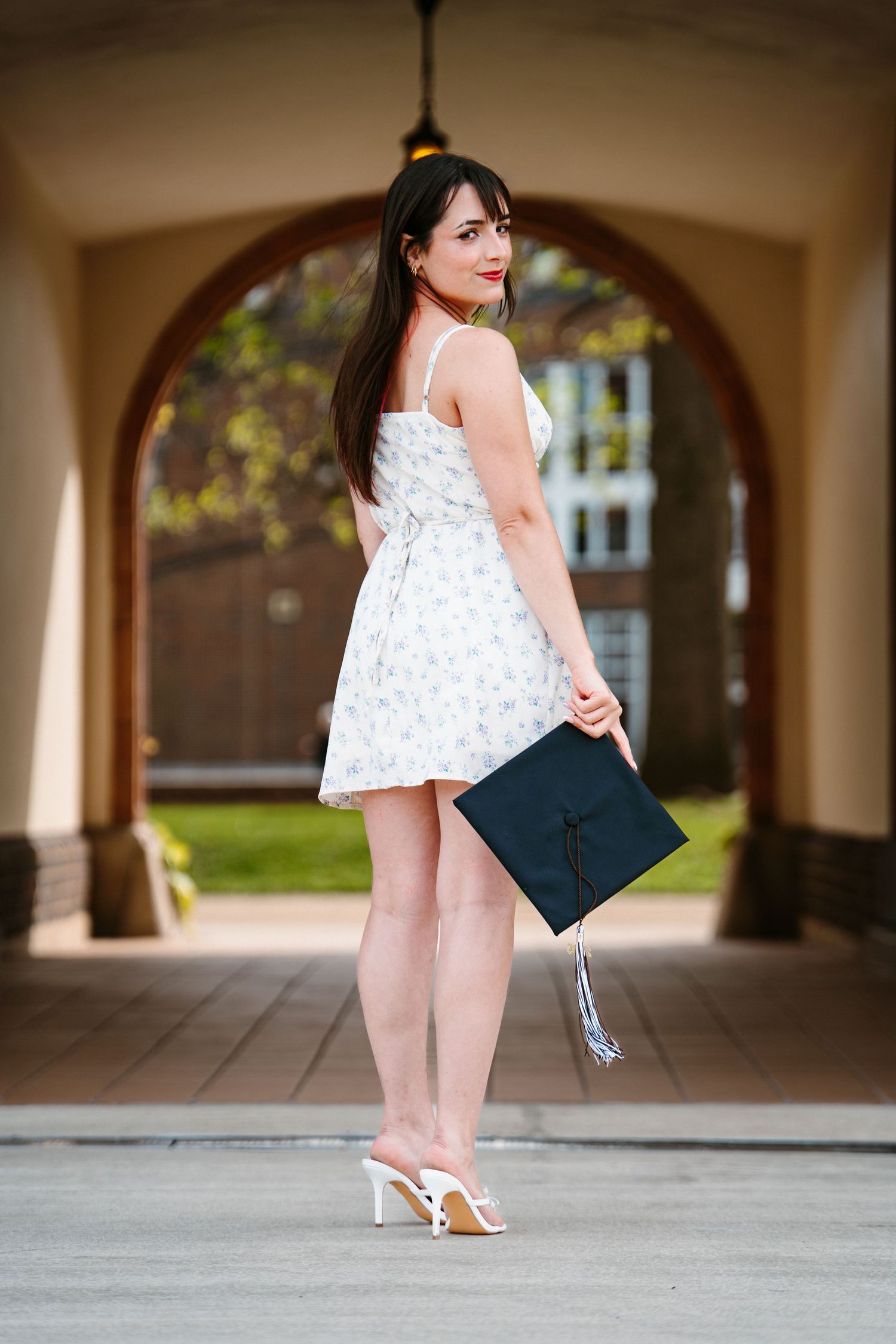 Graduate in a white floral sundress and white heels holding a black mortarboard cap while posing in a sunlit brick archway.