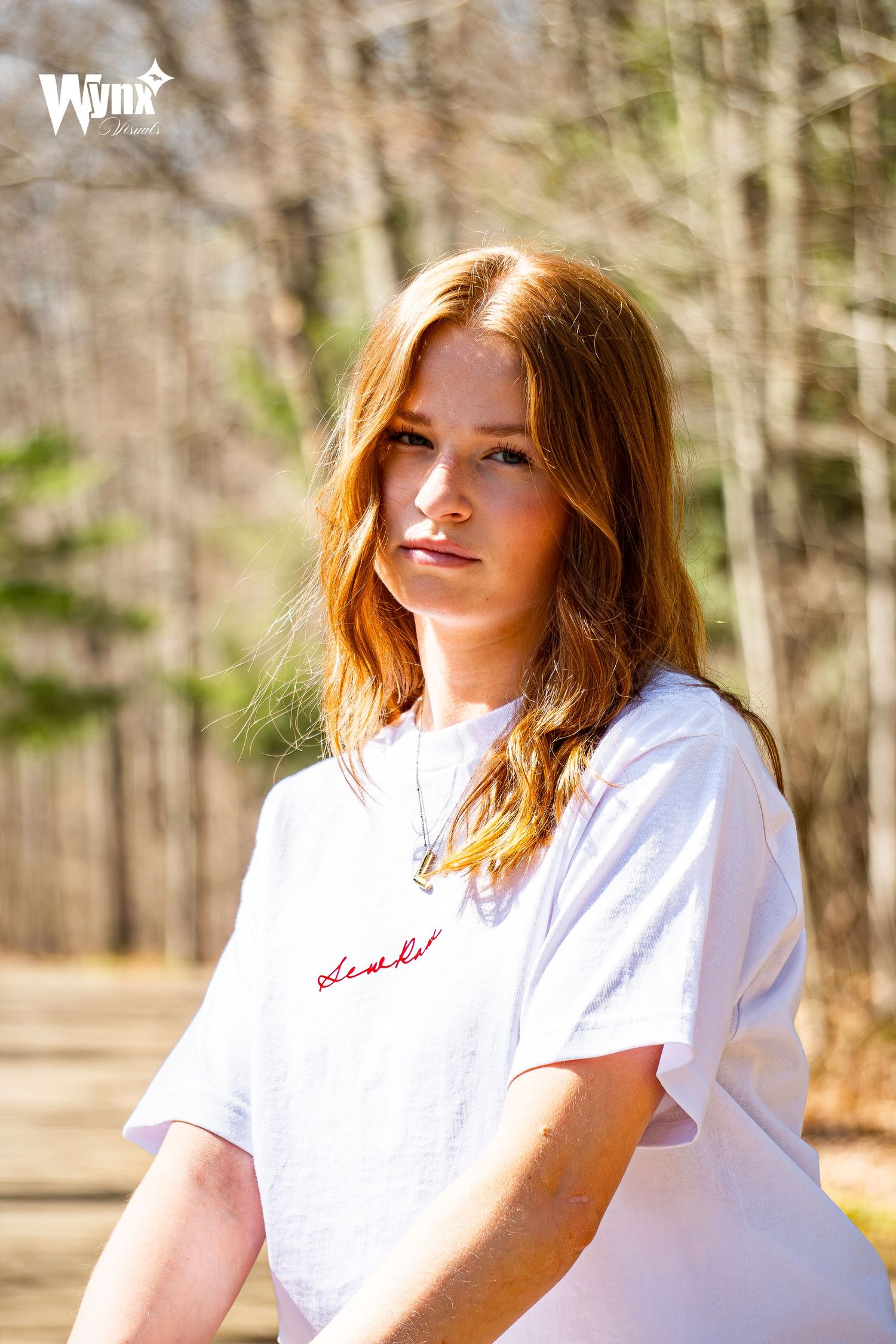 Portrait of a woman with wavy red hair in a white Sewlaw embroidered t-shirt and pendant necklace standing on a sunny trail.