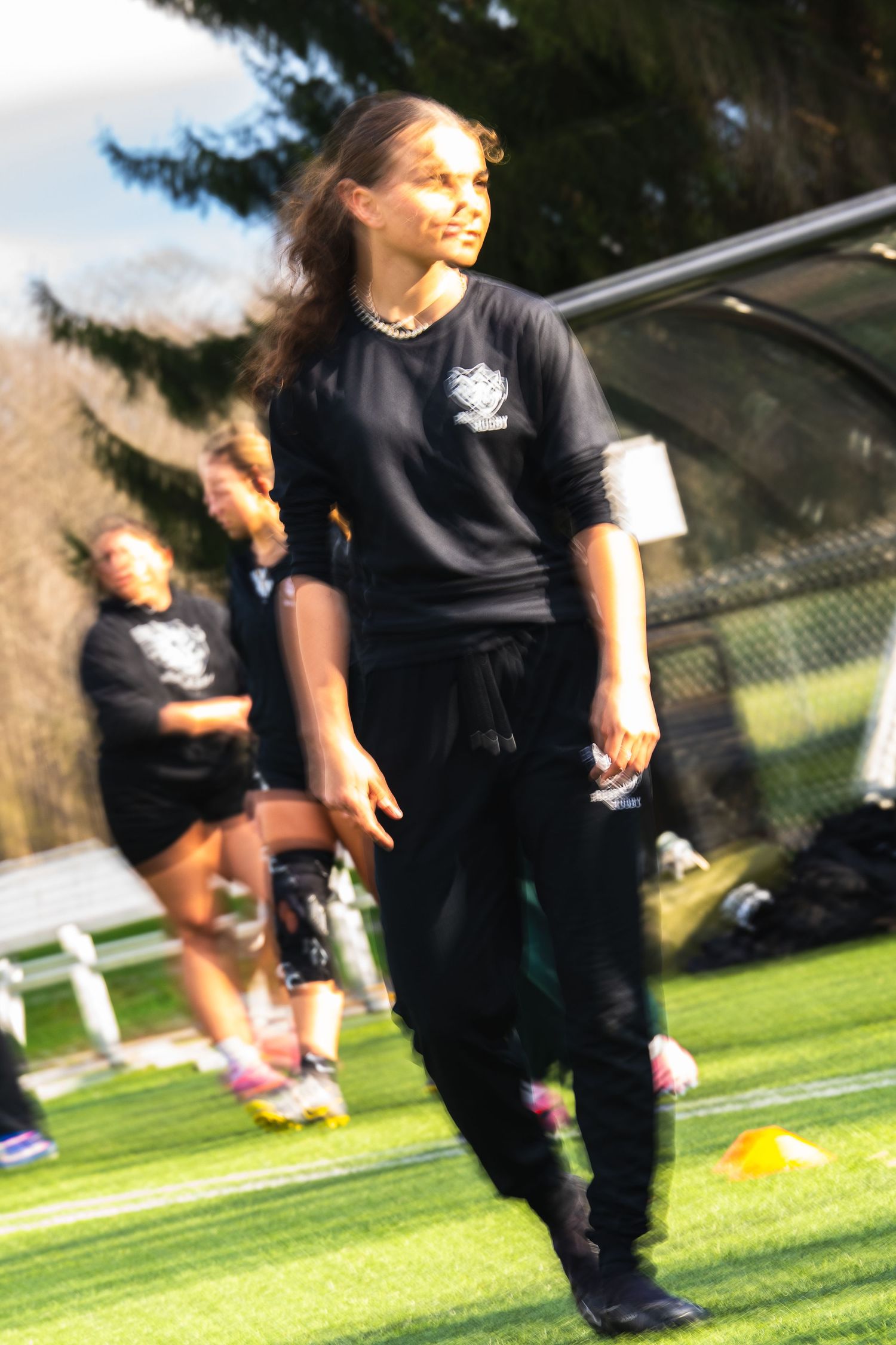 A woman in all-black athletic clothing stands on a turf field looking to the side while teammates warm up behind her.